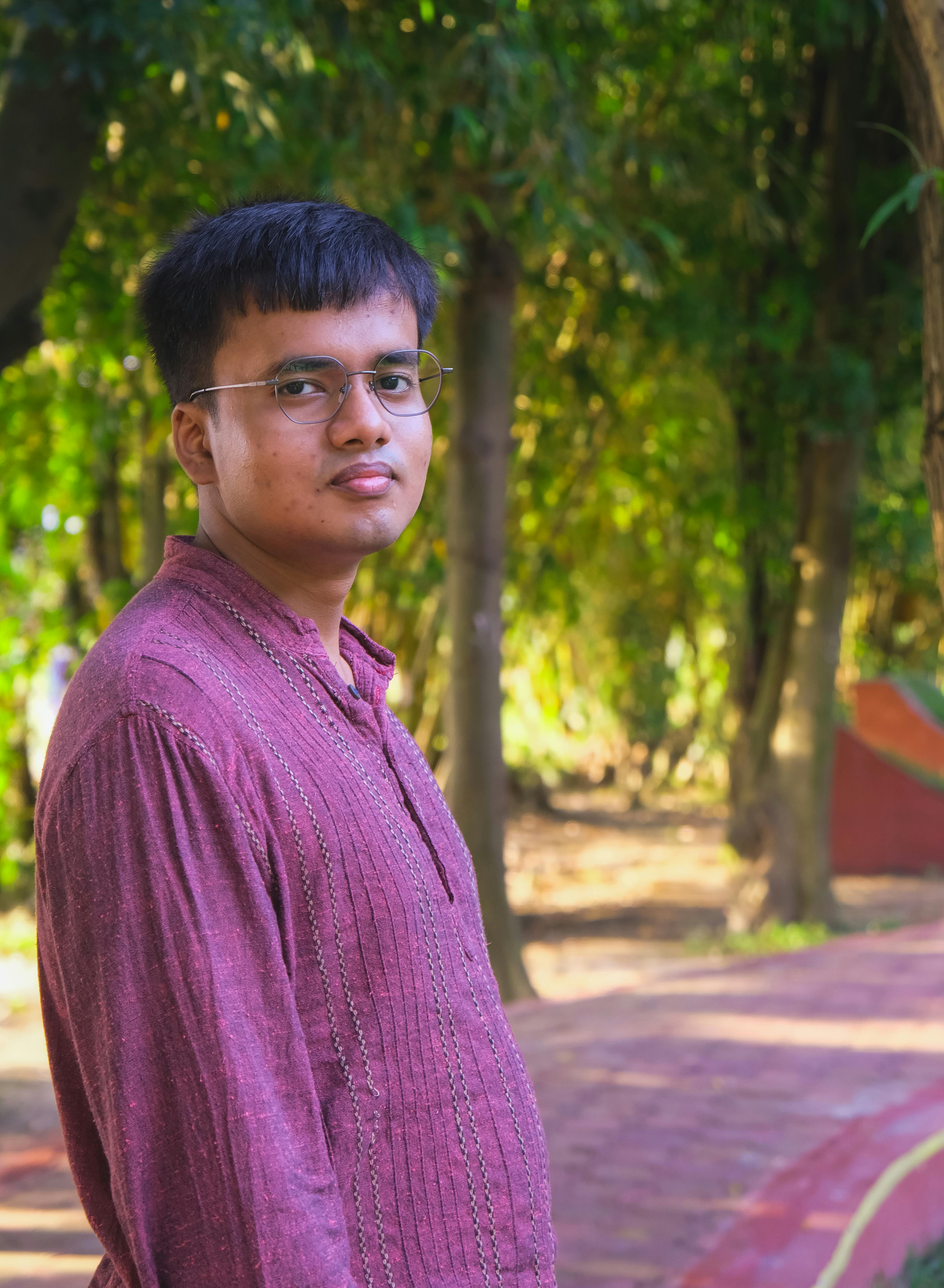 Young man in eyeglasses wearing a pink kurta stands in a serene park setting.
