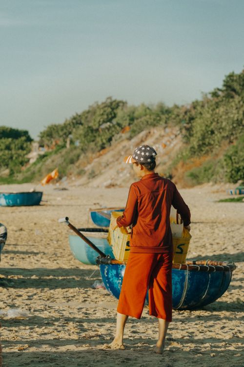 Free Back view of a teenager on a sunny beach carrying a crate, wearing red shorts and sweatshirt. Stock Photo