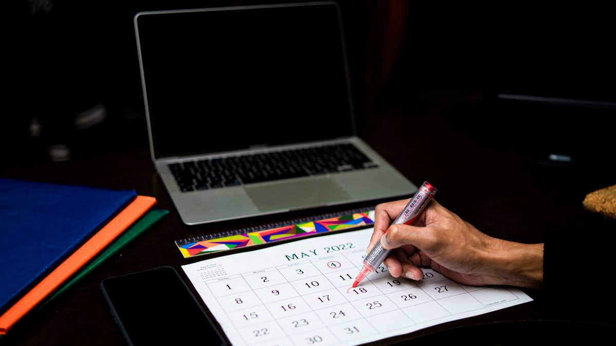 Person planning dates on a desk calendar next to a laptop and phone