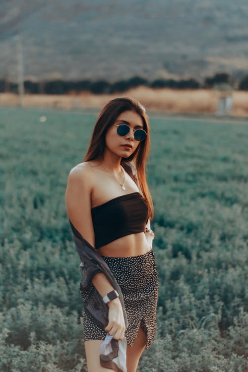 Young Woman in a Summer Outfit and Sunglasses Standing on a Field 
