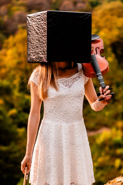 Free Artistic outdoor portrait of a woman in a white dress holding a violin, her head obscured by a box, set against a lush green background. Stock Photo