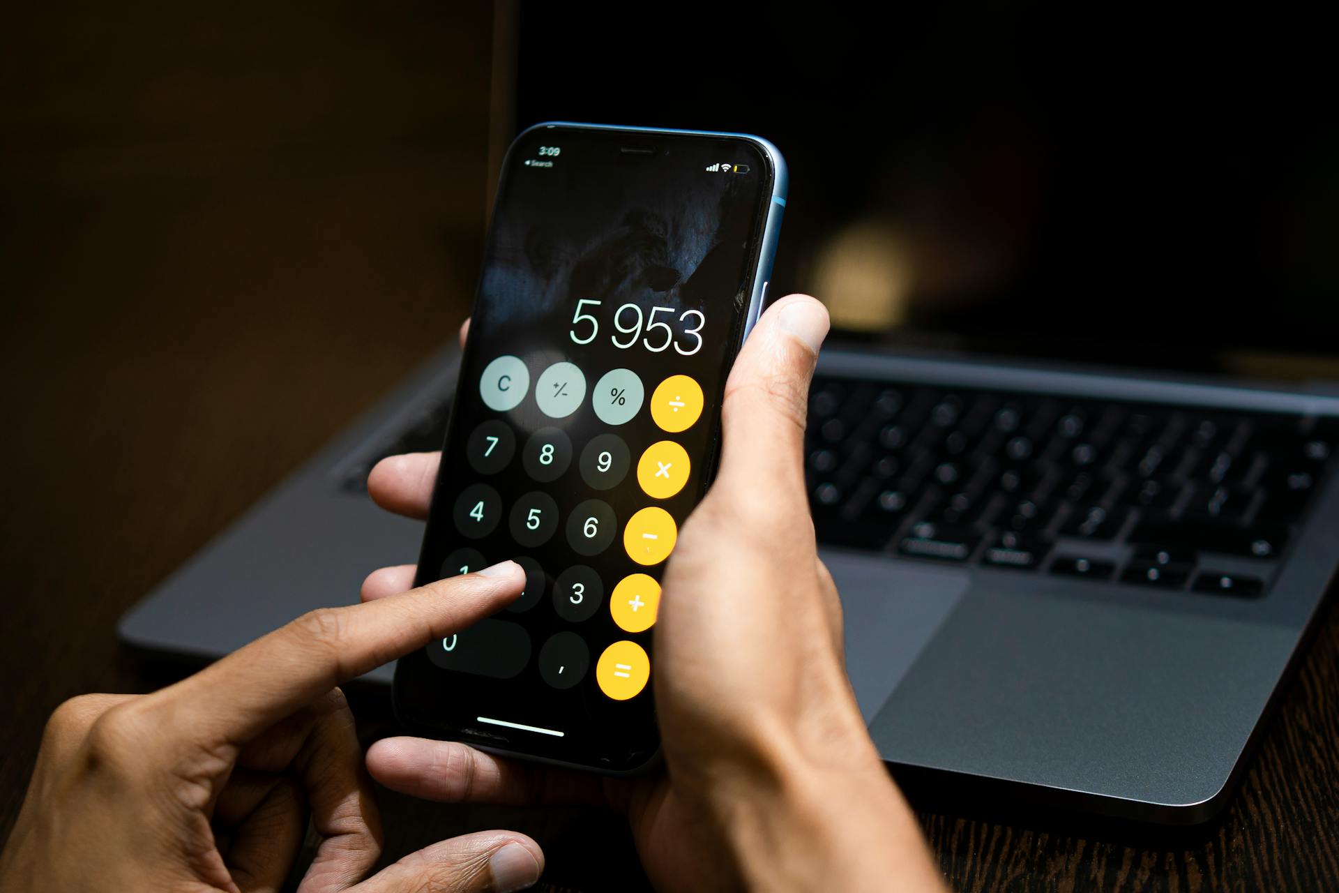 Person using a calculator app on a smartphone with a laptop in the background.