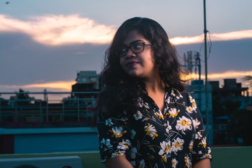 Free A woman in floral attire and eyeglasses poses outdoors at sunset against a city skyline. Stock Photo