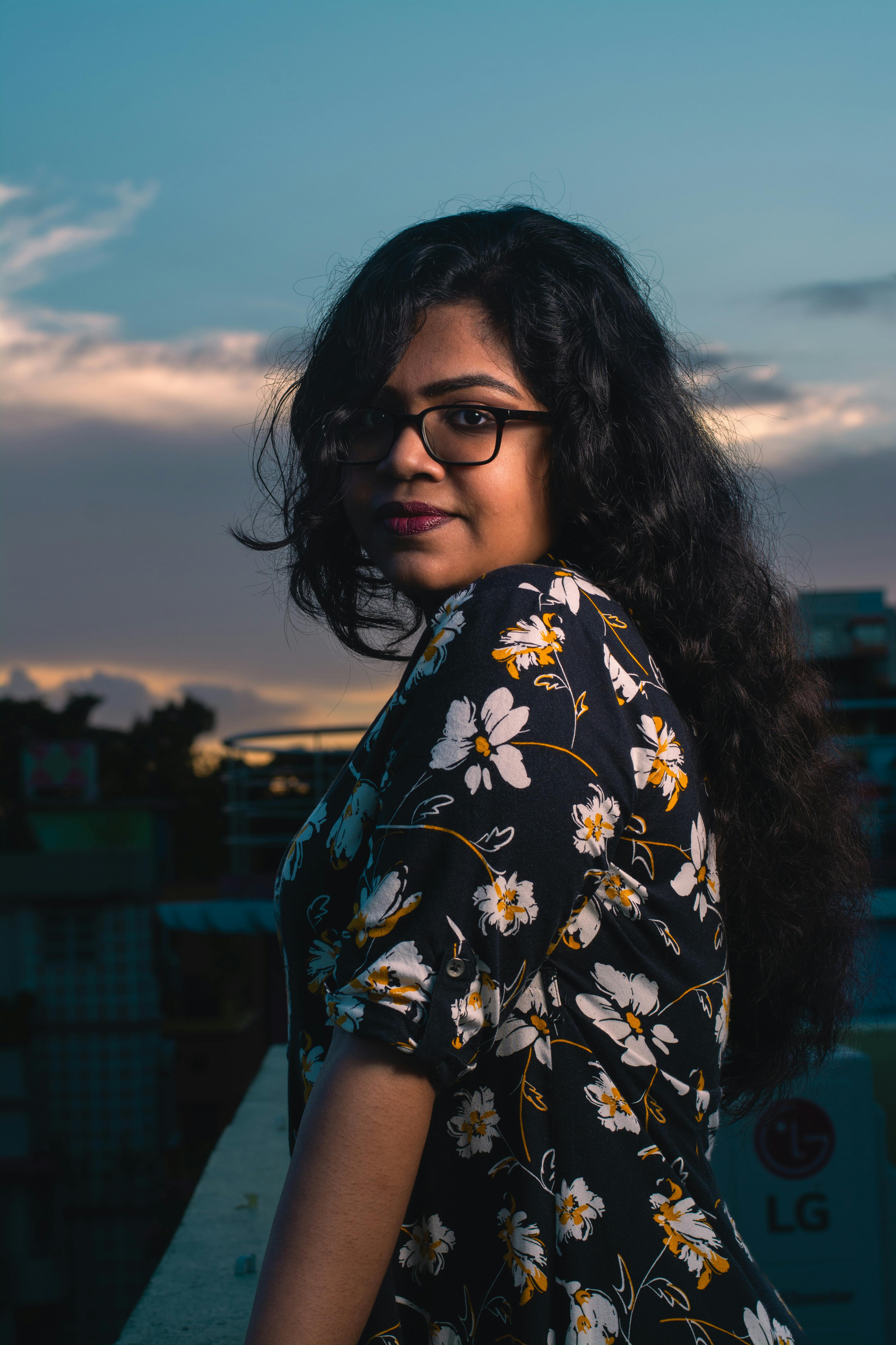 Portrait of a young woman with glasses standing on a city balcony at sunset.
