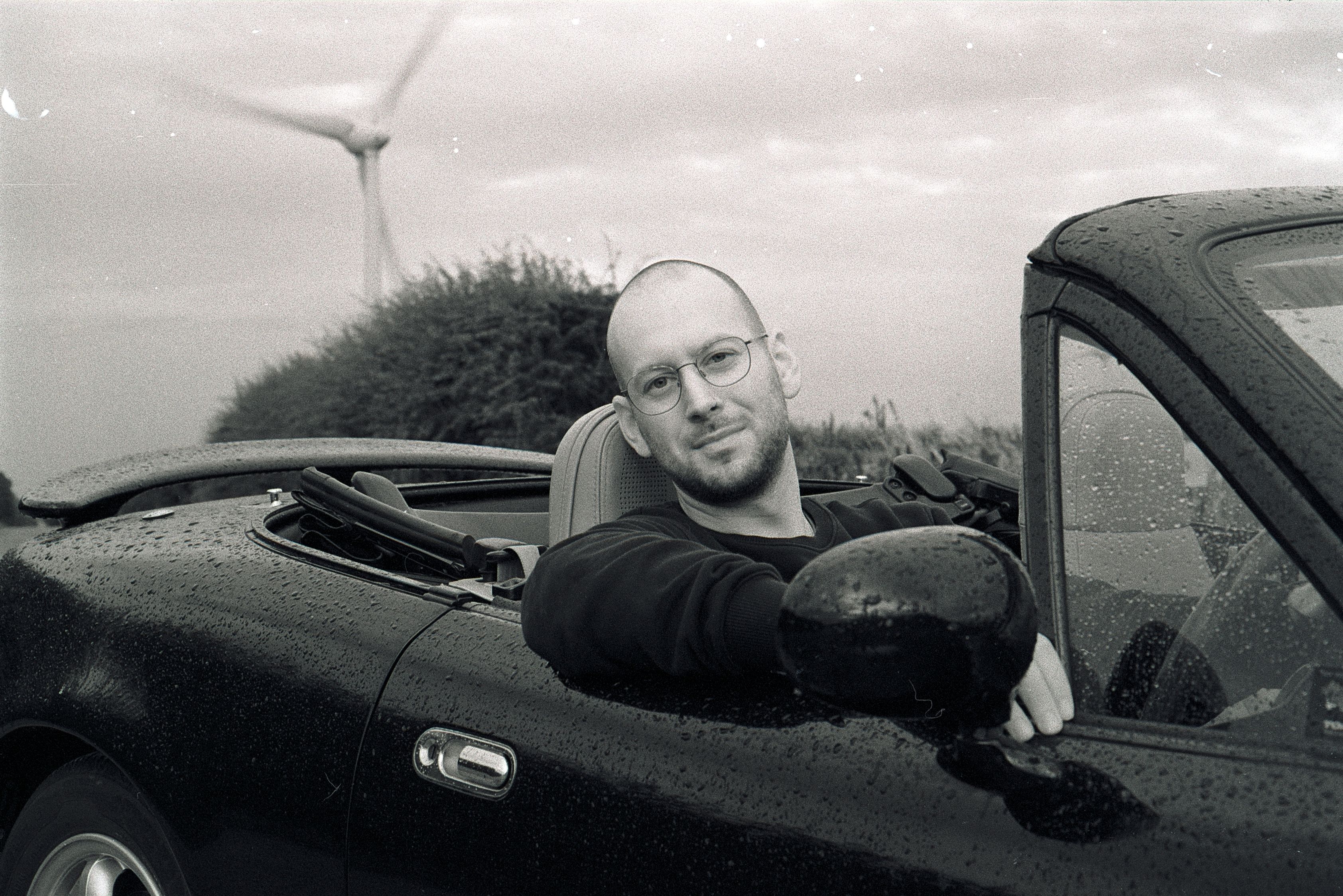 A confident man seated in a convertible car during a rainy day, with a wind turbine in the background.