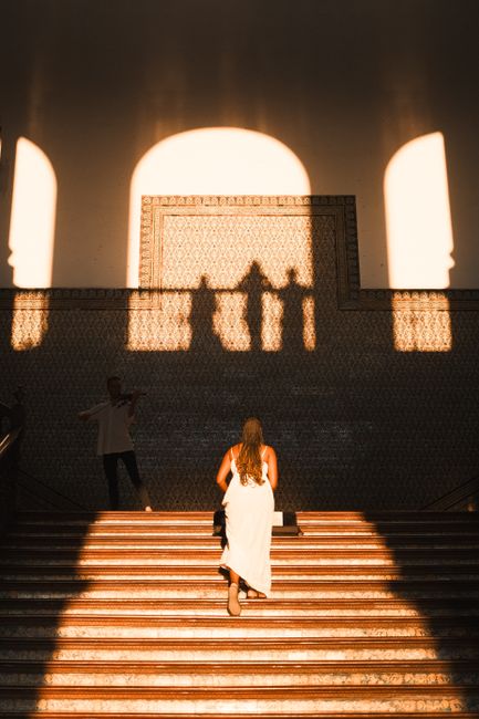 A woman in a dress walks up stairs in Sevilla, casting shadows in warm sunlight.