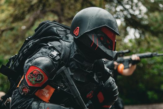 A soldier in advanced tactical armor with a firearm crouches in a forested area during a mission.