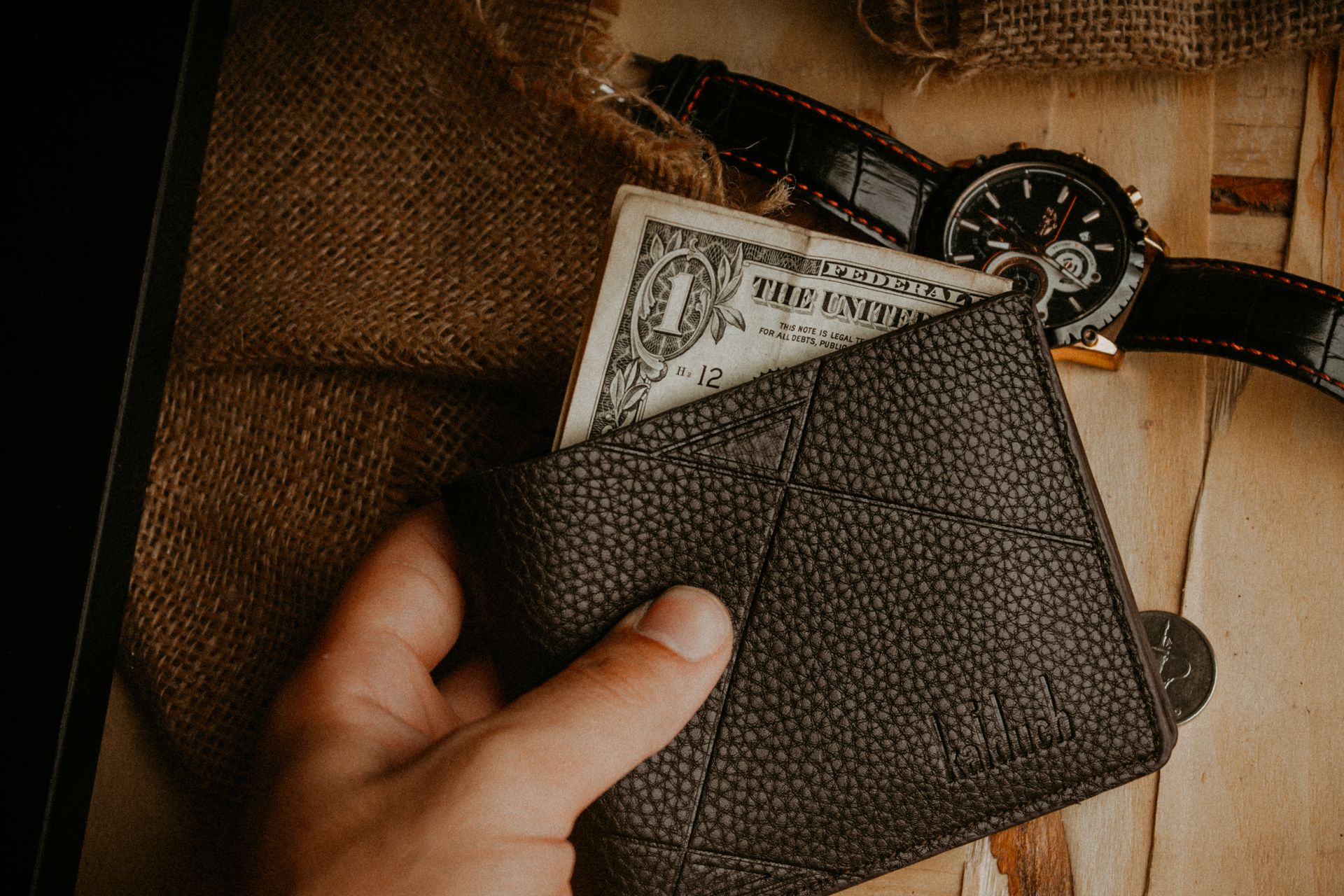 A hand holds a leather wallet with a dollar bill exposed, next to a wristwatch on a wooden surface.