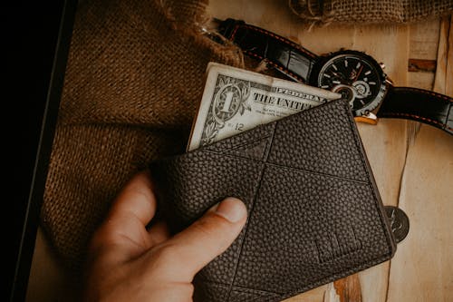 Free A hand holds a leather wallet with a dollar bill exposed, next to a wristwatch on a wooden surface. Stock Photo
