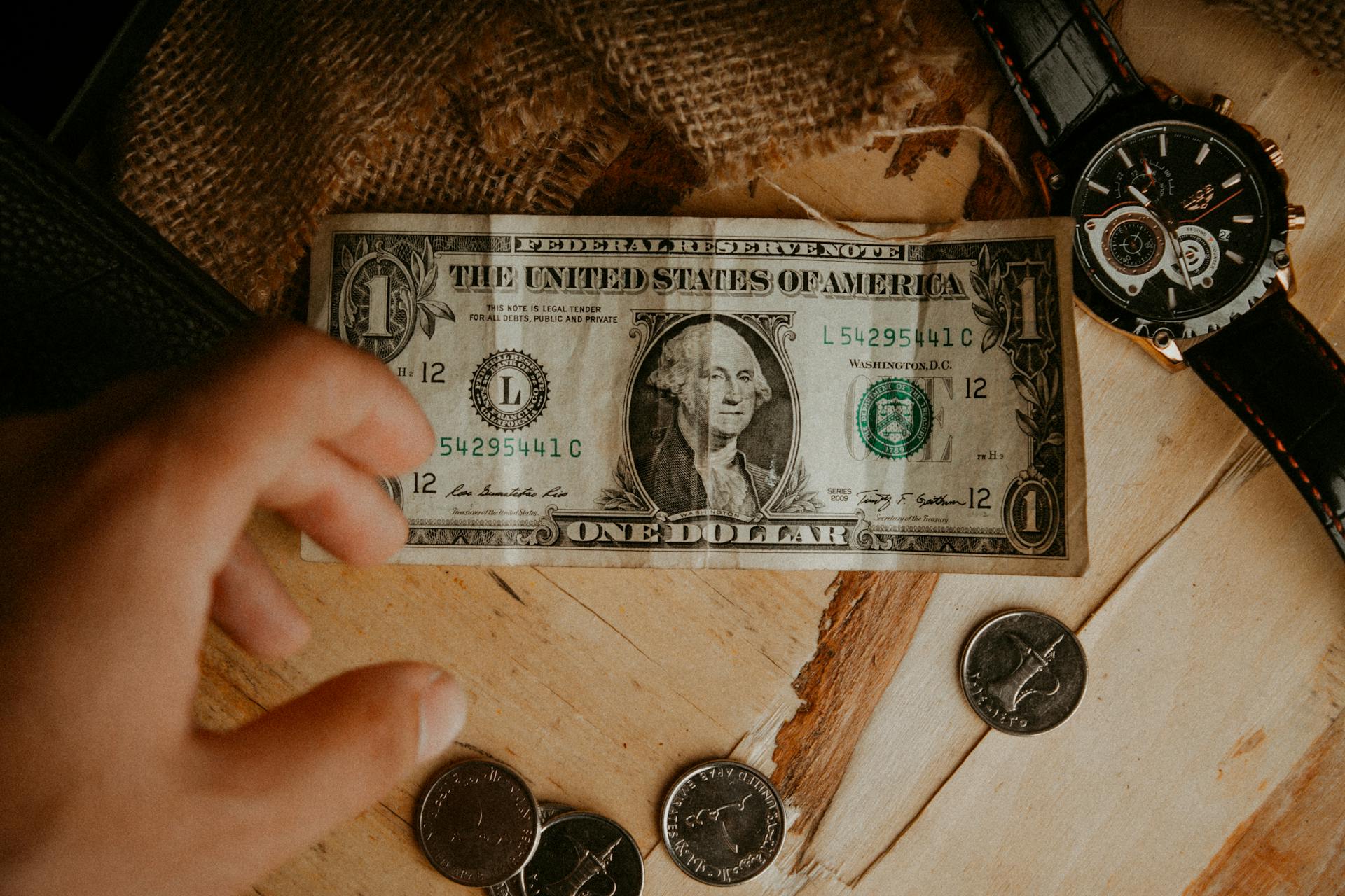 Close-up of a hand reaching for a dollar bill and coins on a wooden table with a watch.