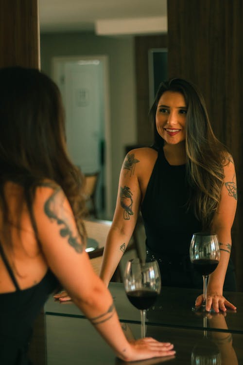 Young Woman with Tattoos Standing in front of the Mirror with a Glass of Wine and Smiling 