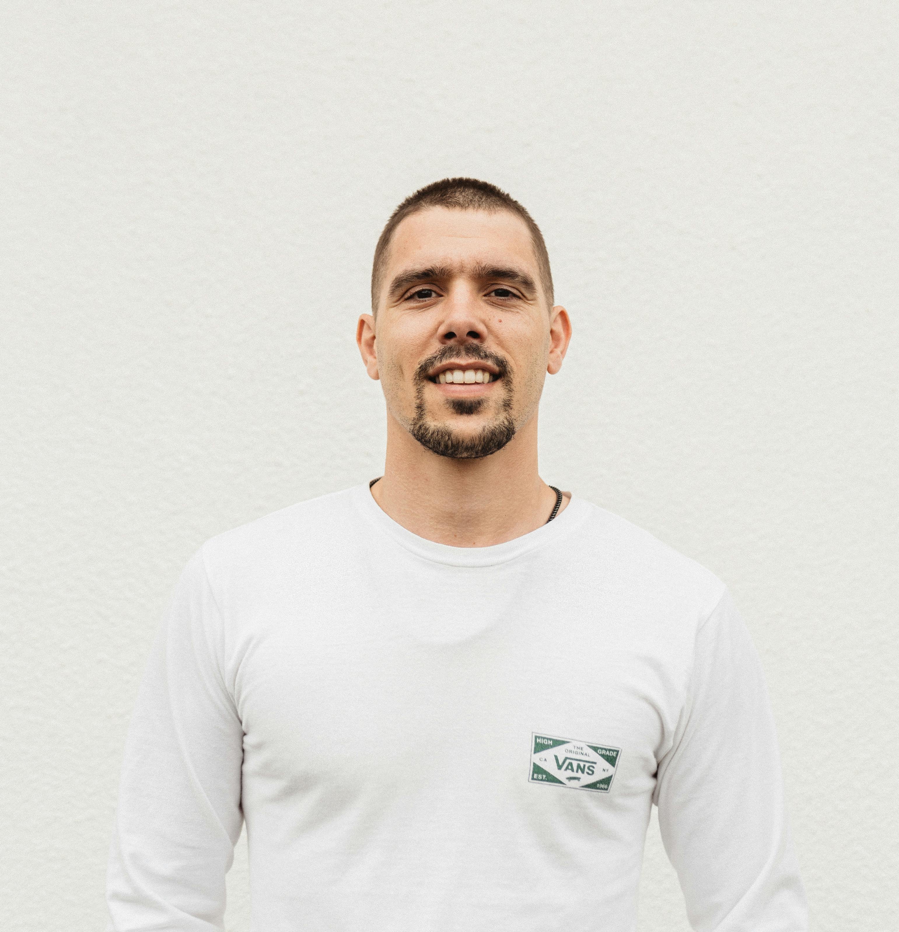 Portrait of a smiling man in a white shirt standing against a white wall with a neutral expression.