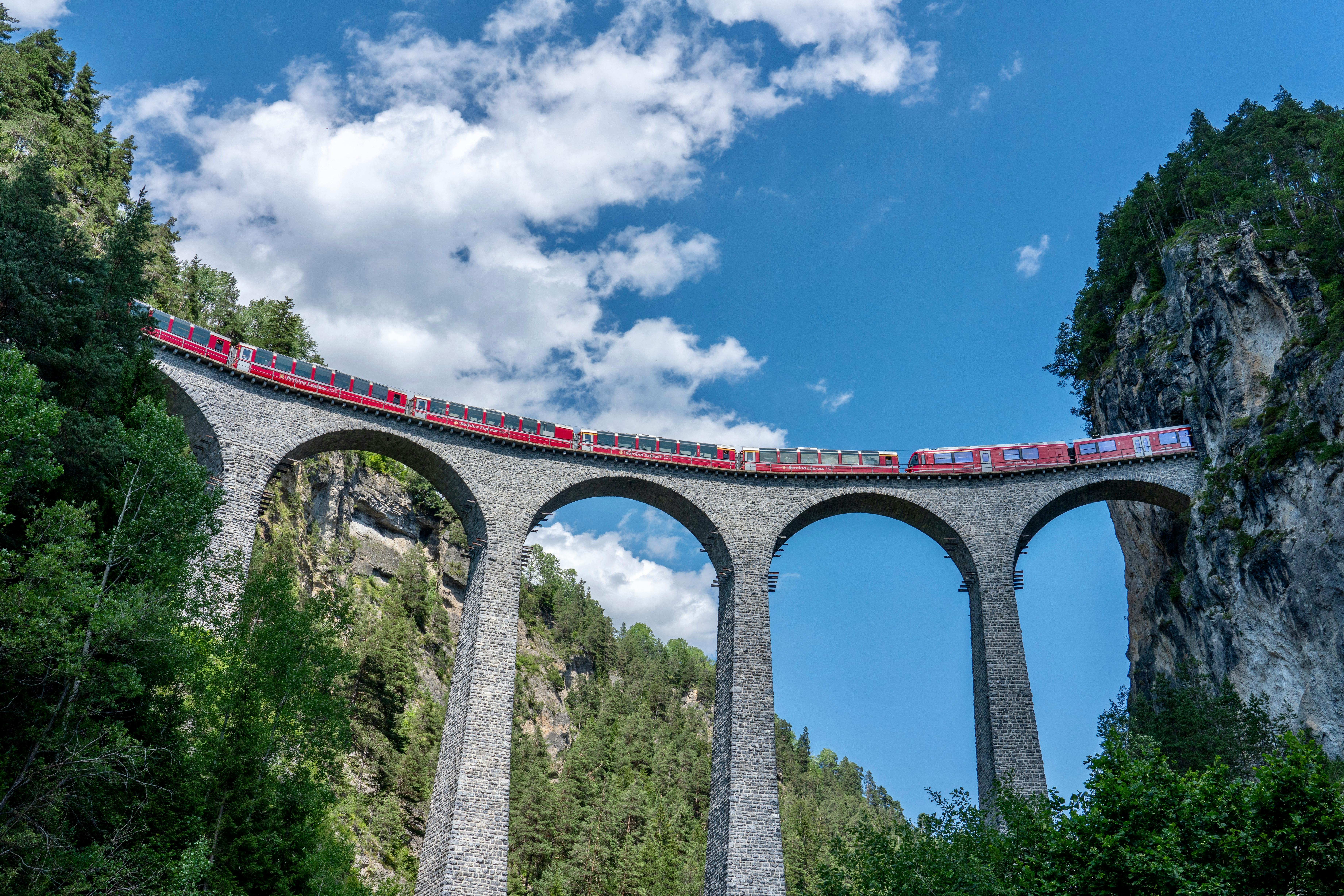 Red train crosses the iconic Landwasser Viaduct in Switzerland, surrounded by stunning alpine scenery.