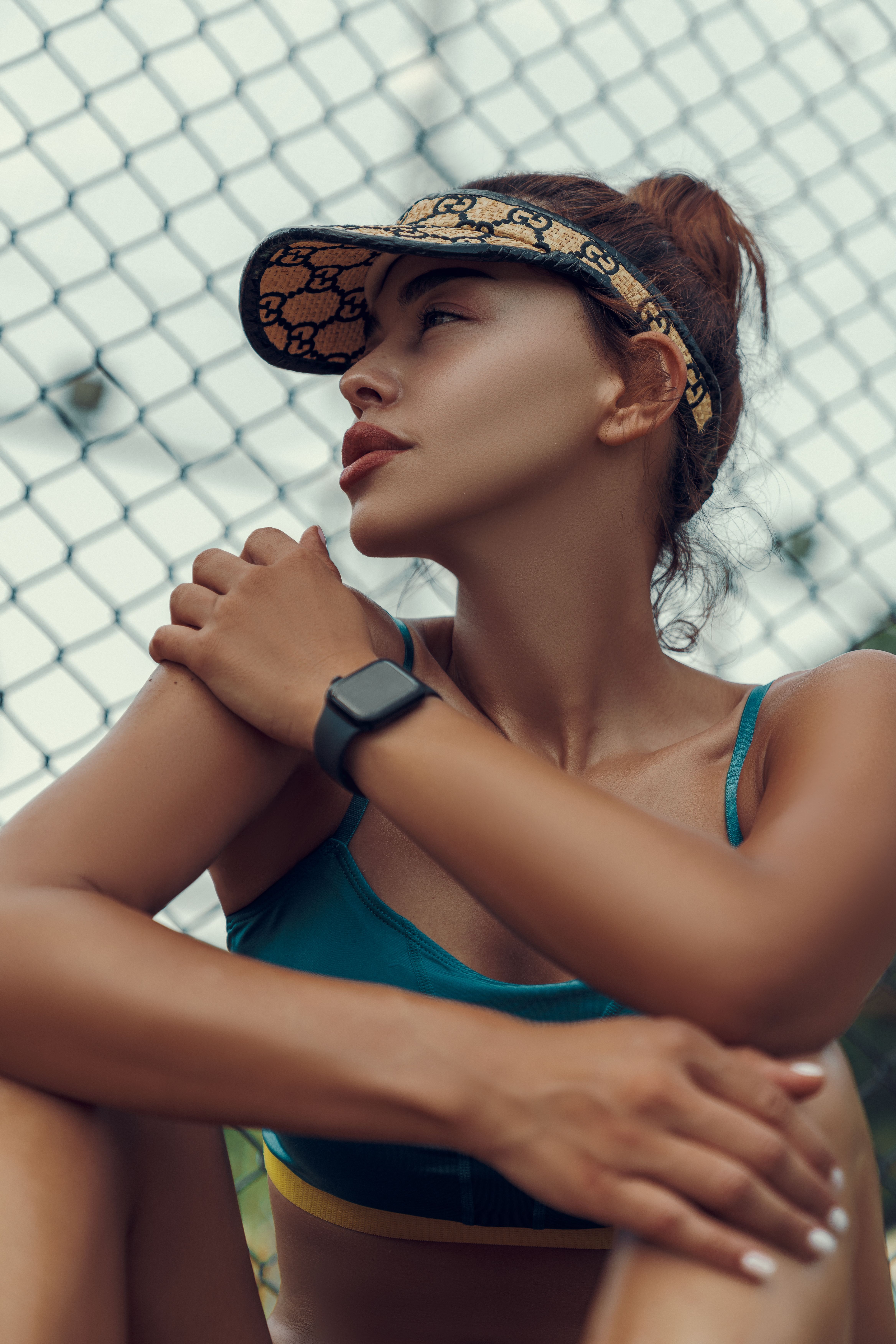 Young woman in sportswear sits by a fence wearing a sun visor, exuding confidence.
