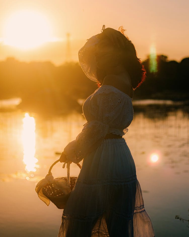 Woman In Dress Standing By A Body Of Water At Sunset 