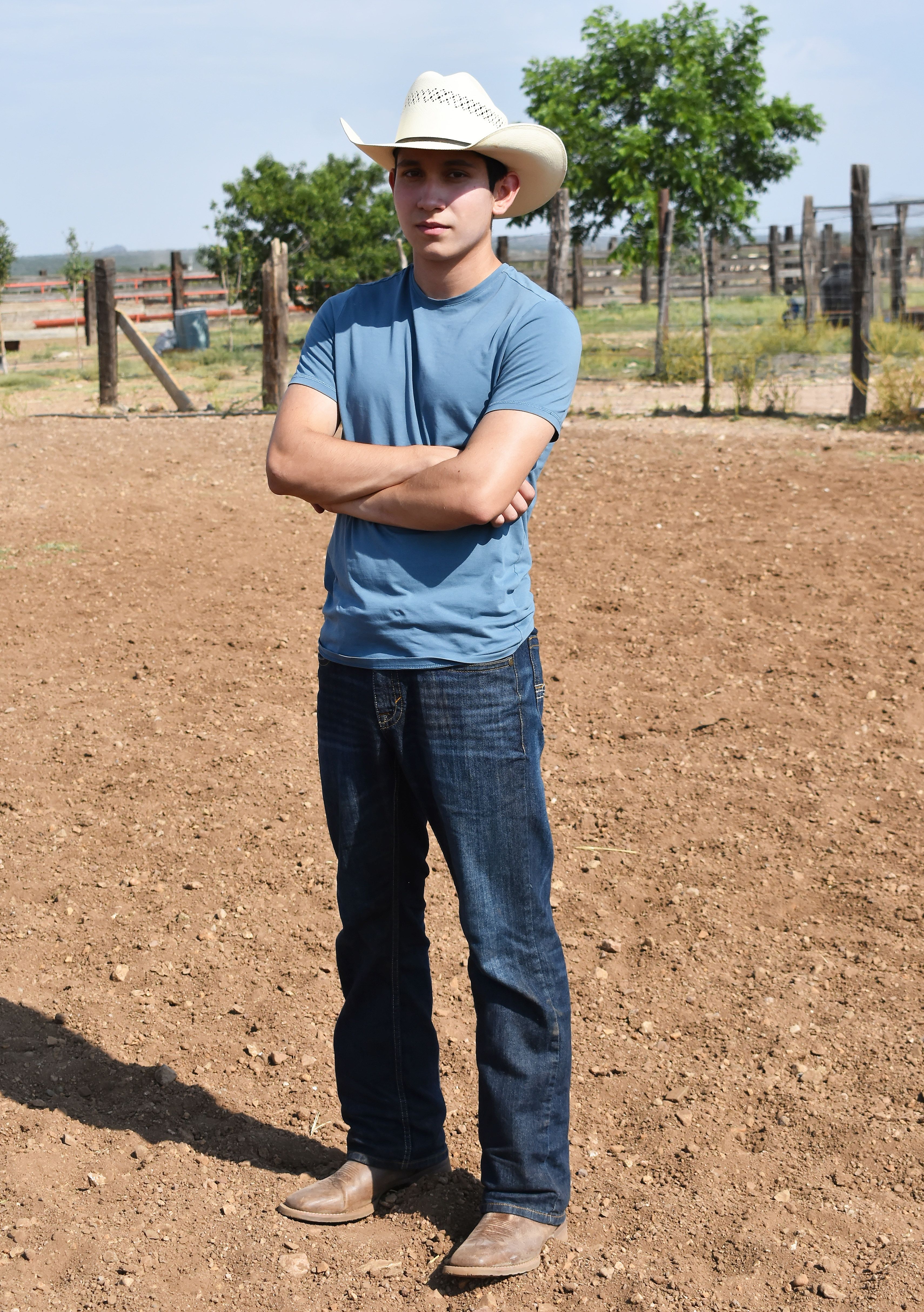 Young man in cowboy hat standing confidently on a farm under a sunny sky.