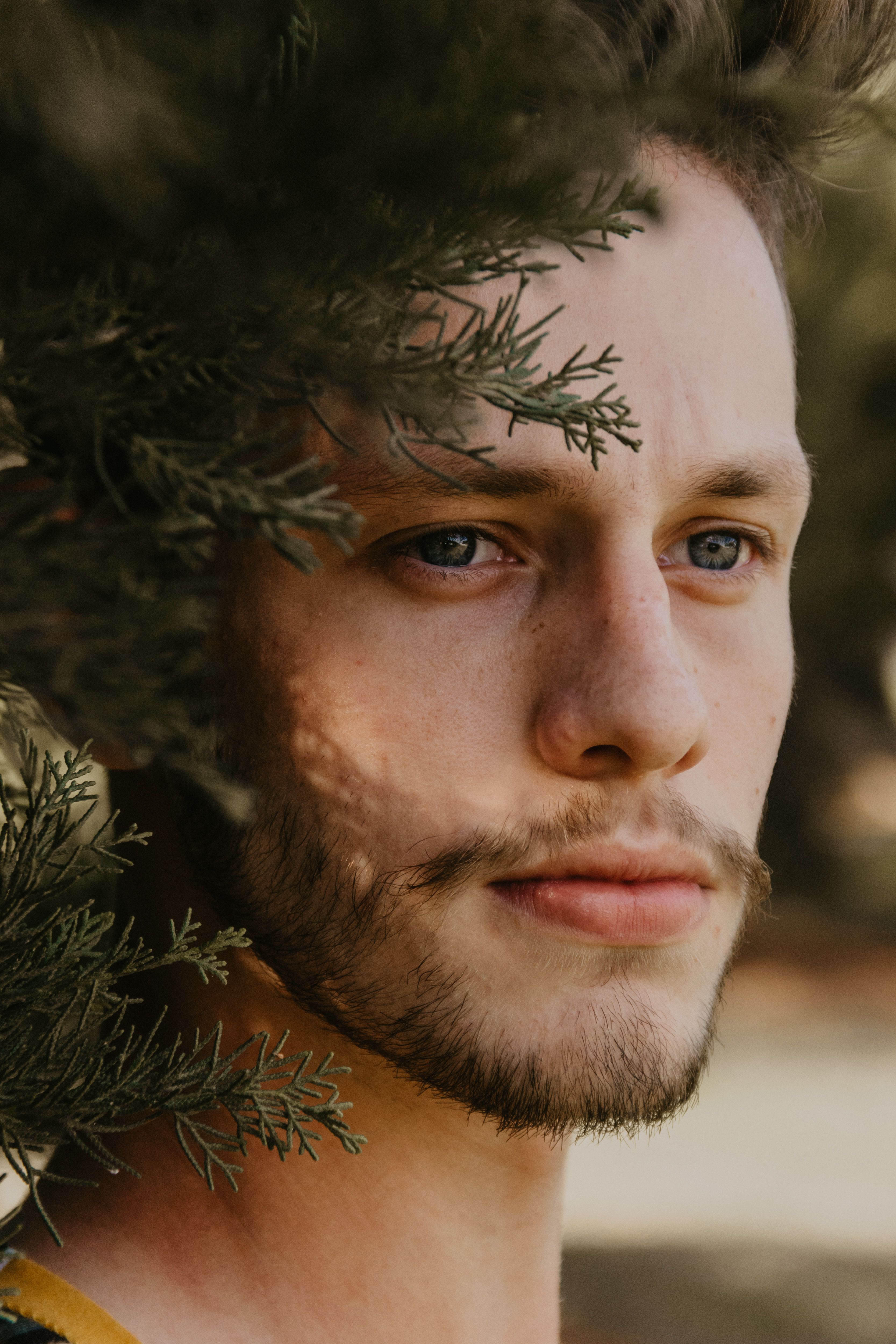 Close-up portrait of a young man with stubble framed by conifer branches.
