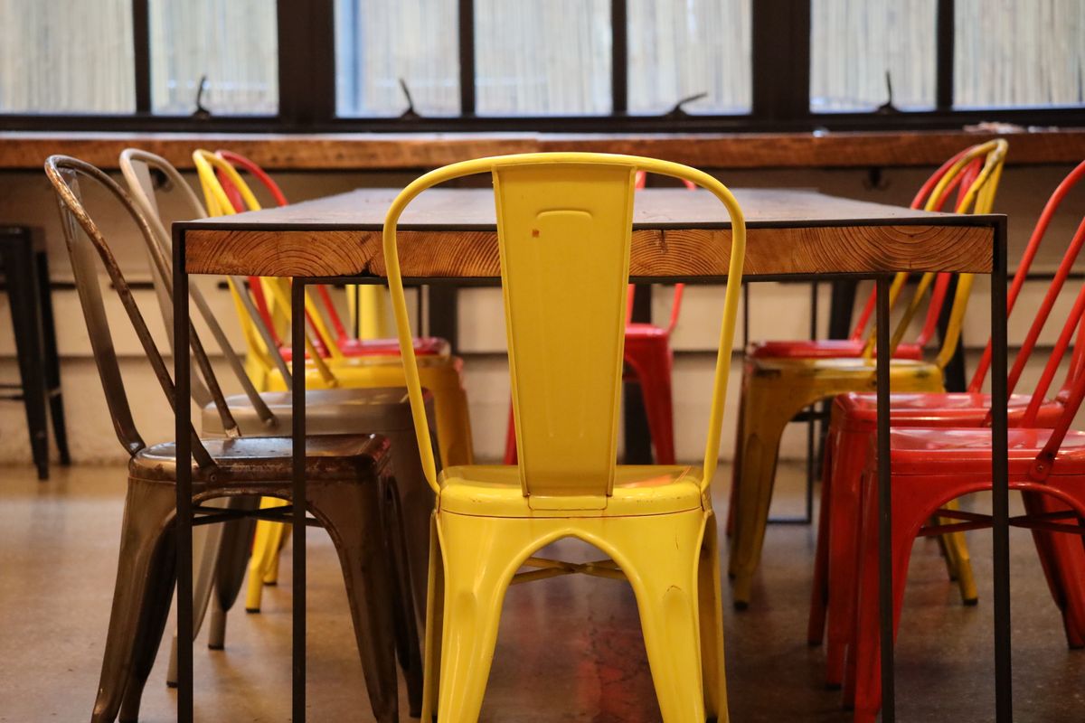 Scaune industriale retro în culori vii, așezate în jurul unei mese din lemn într-un spațiu interior de tip loft