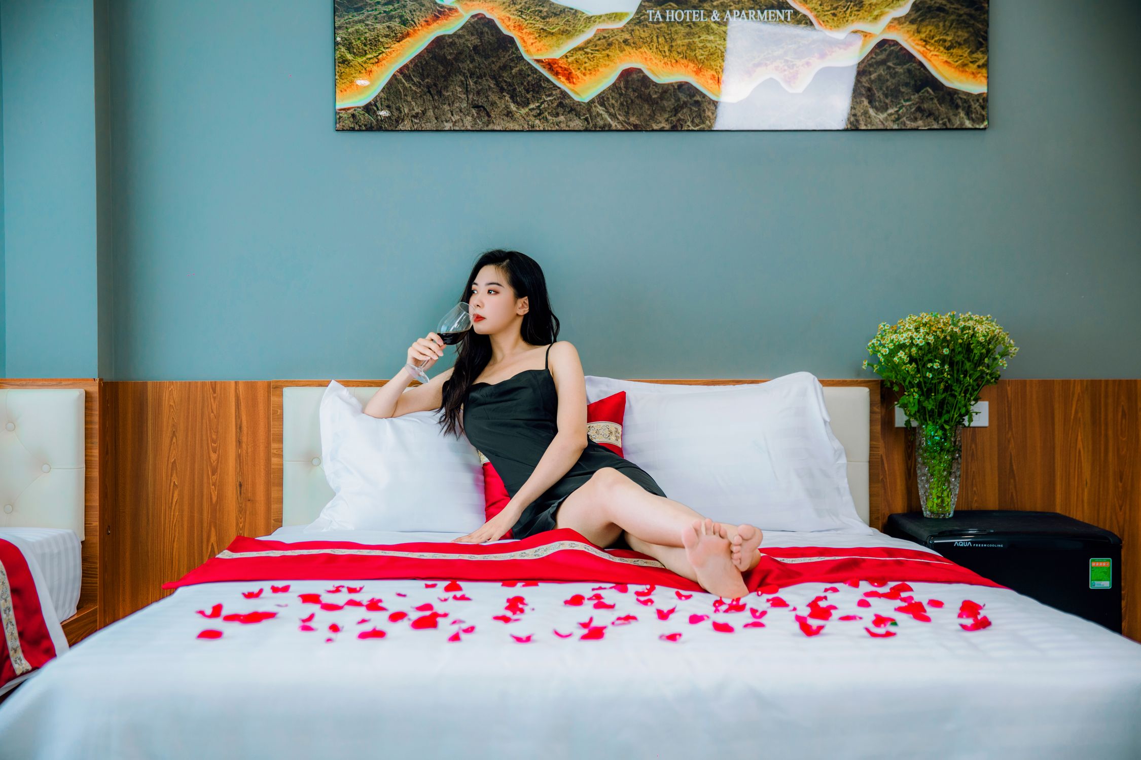 A woman lounges on a bed with rose petals, sipping wine in a chic bedroom setting.
