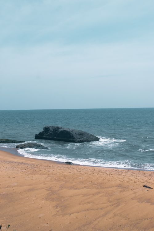 Free Peaceful beach scene with gentle waves, sandy shore, and rocky outcrop under a clear sky. Stock Photo