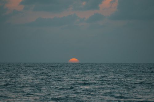 Free A tranquil view of the sun setting over the calm ocean horizon with soft cloud cover. Stock Photo