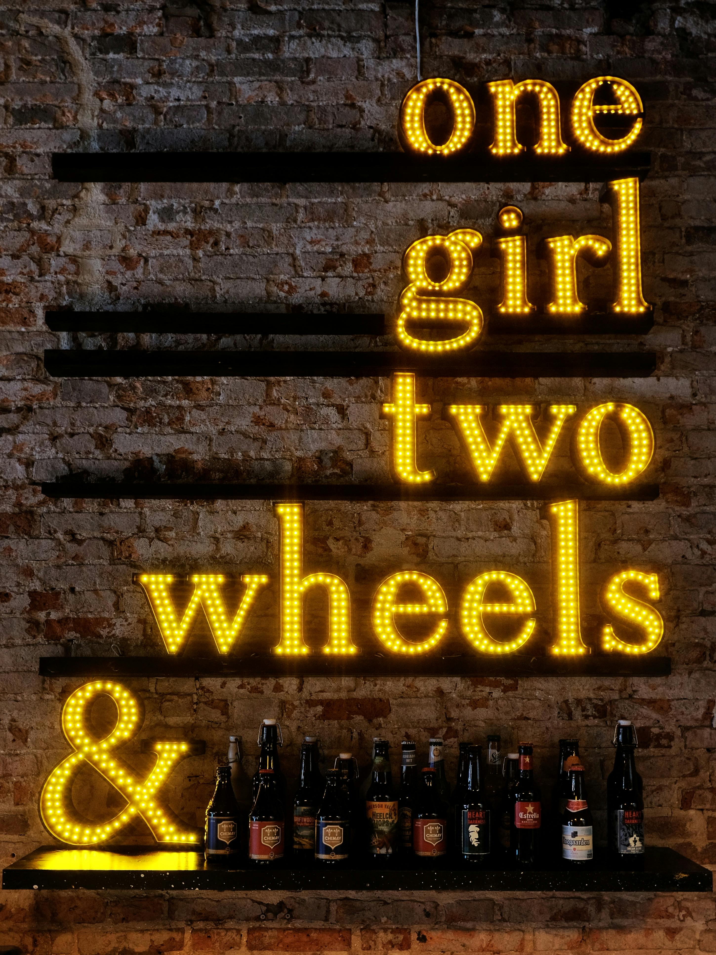Neon styled bar sign reading 'one girl two wheels &' with a row of bottles underneath on a brick wall.