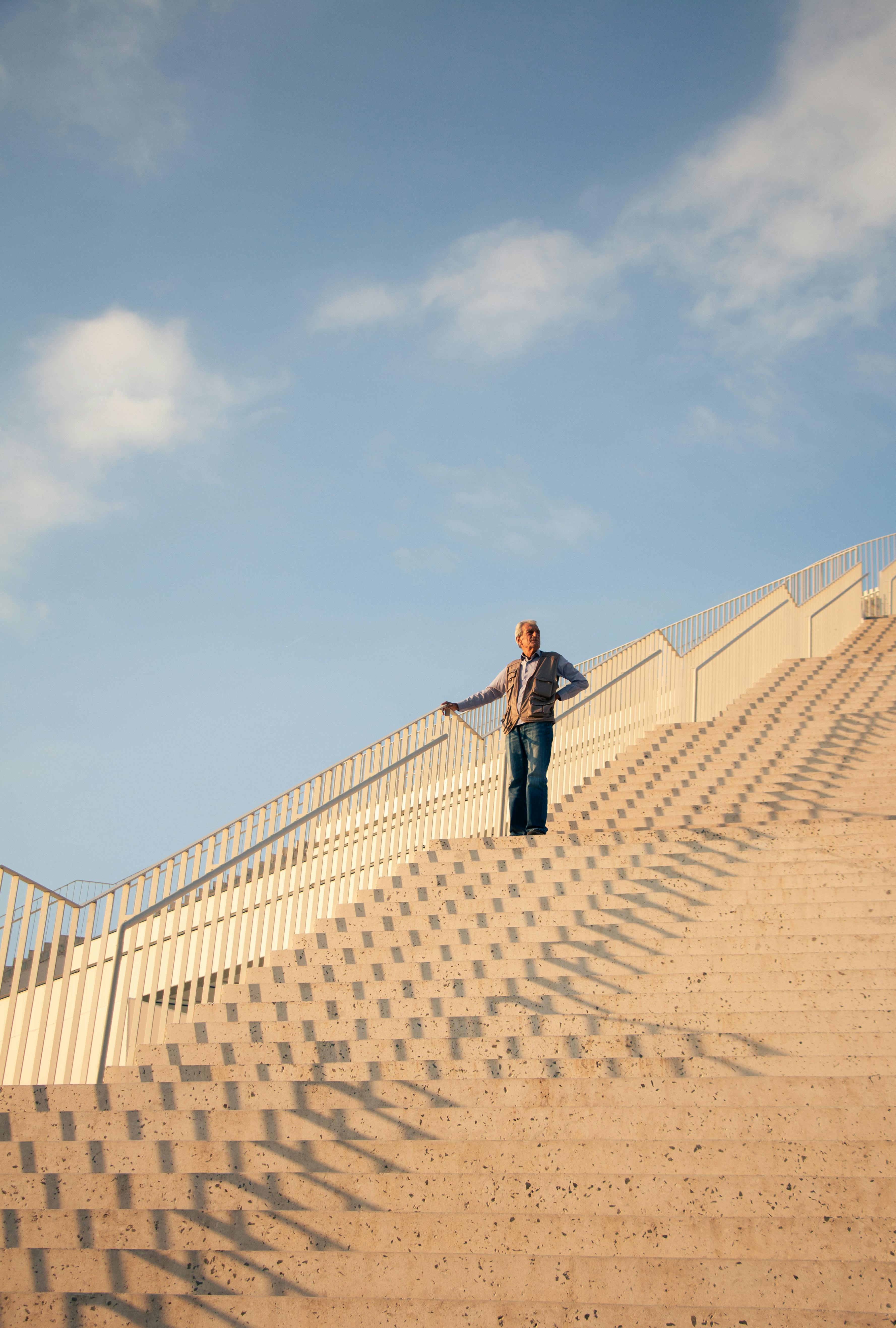 An elderly man stands on a sunlit staircase, casting a shadow against a clear blue sky in Tirana.