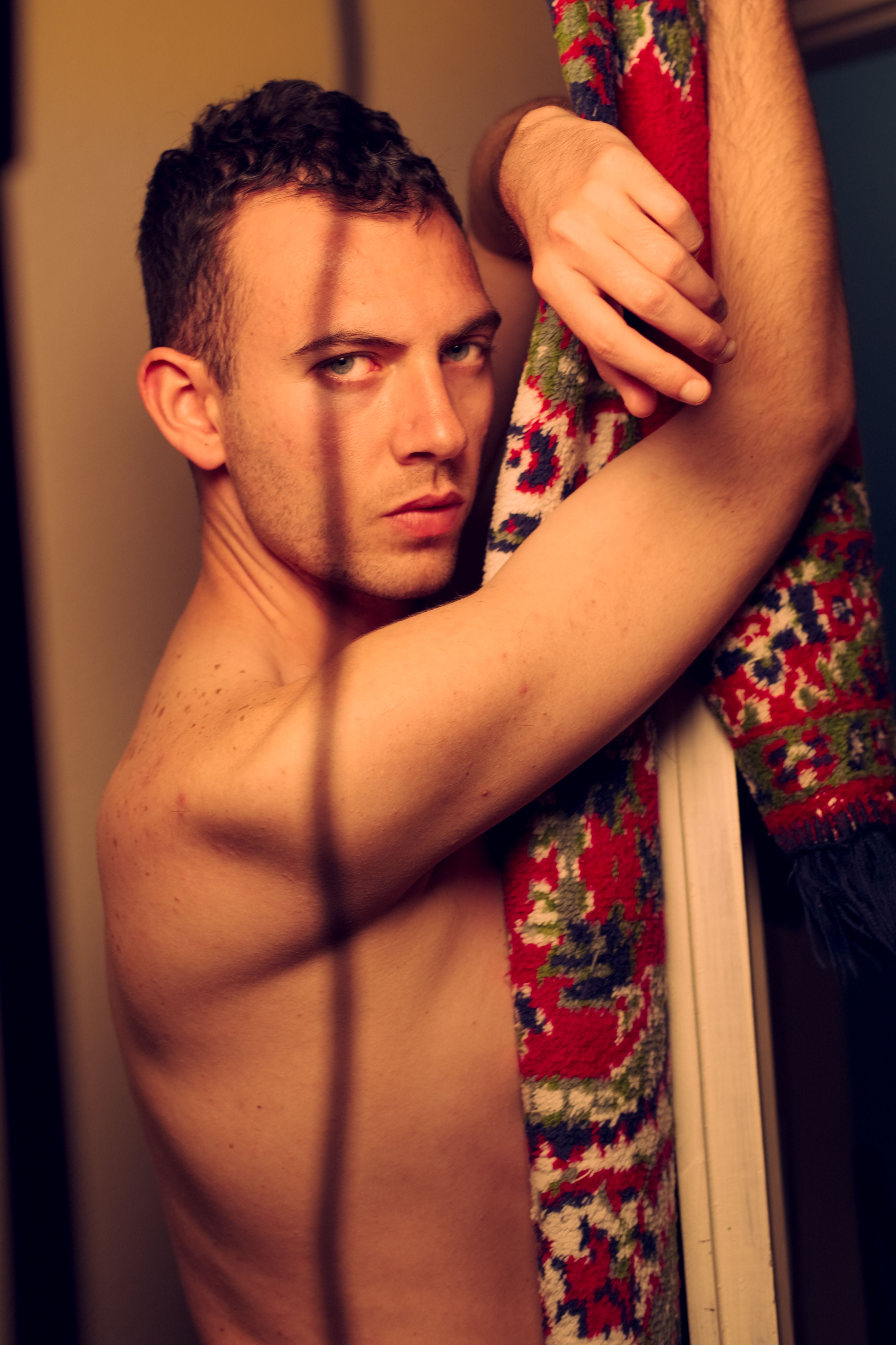 Dramatic portrait of a shirtless man posing indoors next to a vibrant patterned carpet.