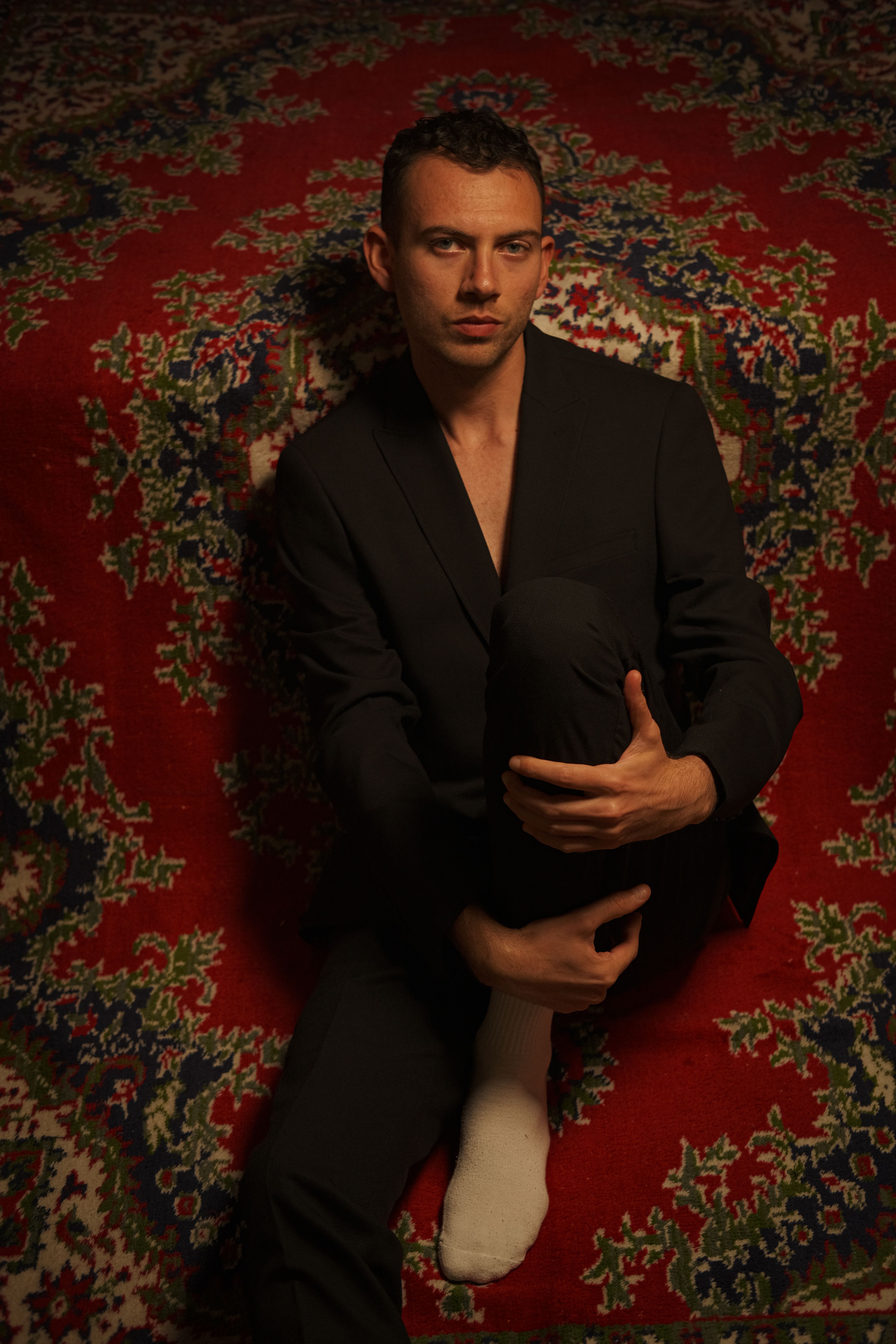 Fashion portrait of a young man in a blazer sitting on an ornate red carpet.