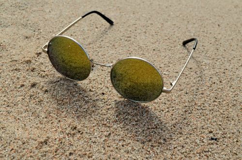 Free Close-up of round sunglasses lying on a sandy beach with a retro fashion vibe. Stock Photo
