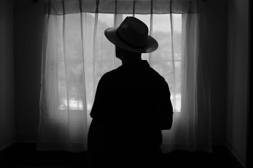 Silhouette of a Man Wearing a Hat against a Window with a Curtain