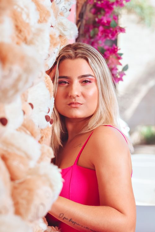 Free Blonde woman in pink stands beside teddy bears and vibrant flowers in a sunny outdoor setting. Stock Photo