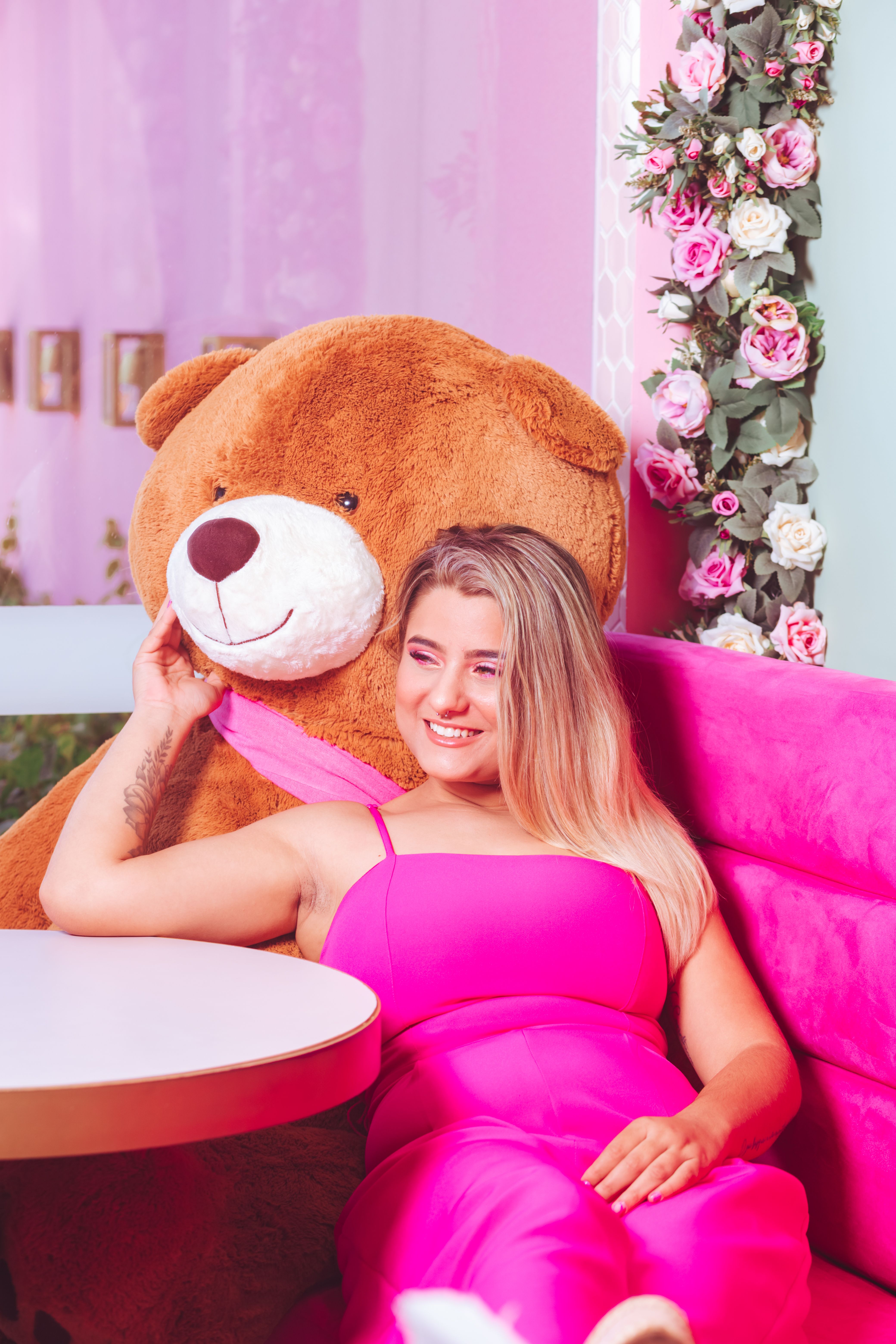 Smiling woman in pink dress relaxing with a large teddy bear in a vibrant pink-themed cafe.