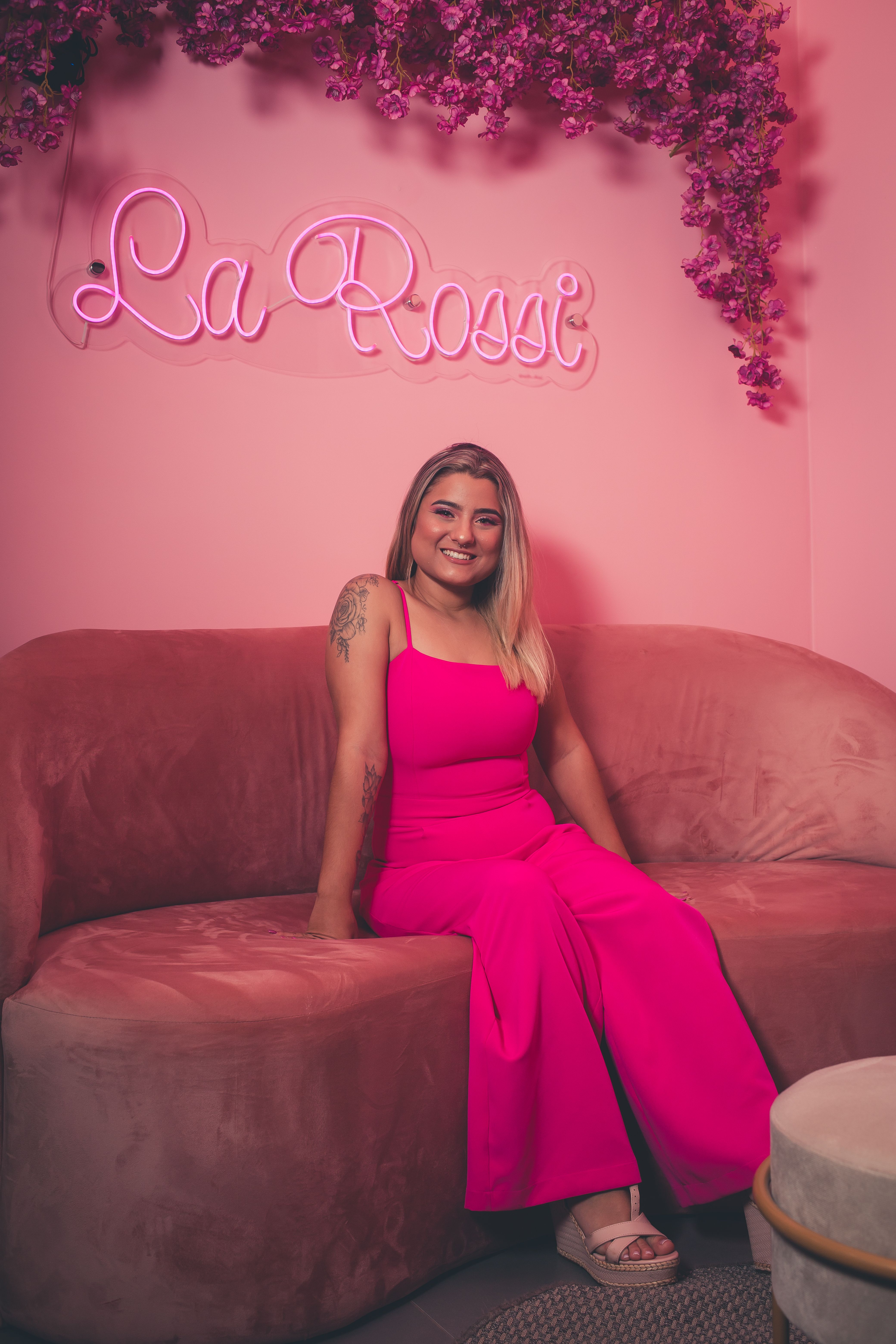Smiling woman in a bright pink outfit sitting on a plush sofa under neon signage.