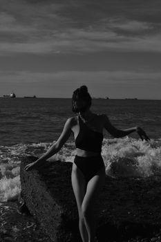Young woman posing in black swimsuit on a wave breaker at the beach, captured in black and white.