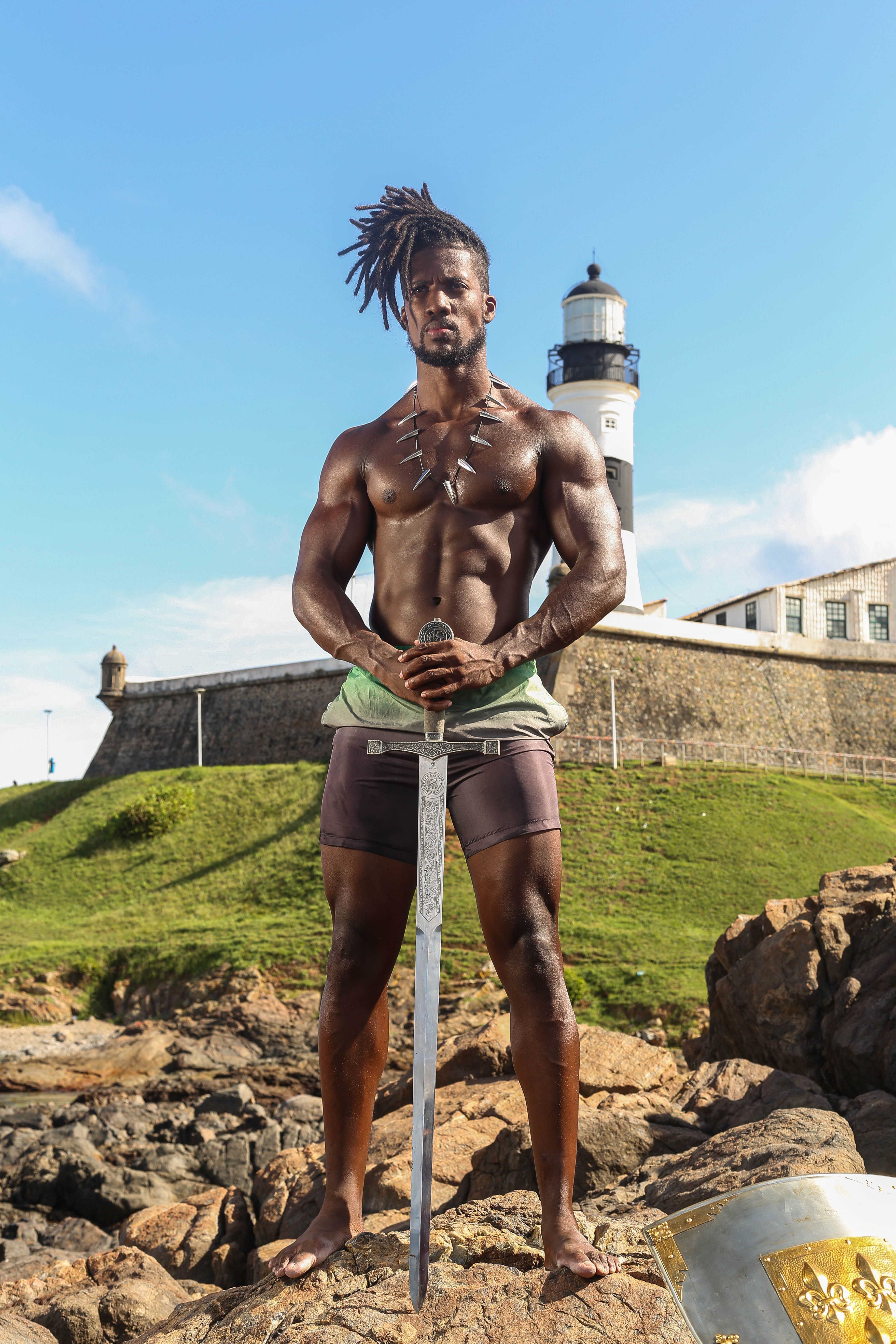 Fit black man stands confidently with sword near Salvador lighthouse on rocks.