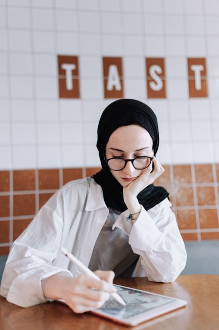 Focused woman in glasses and hijab works on tablet in stylish cafe setting.