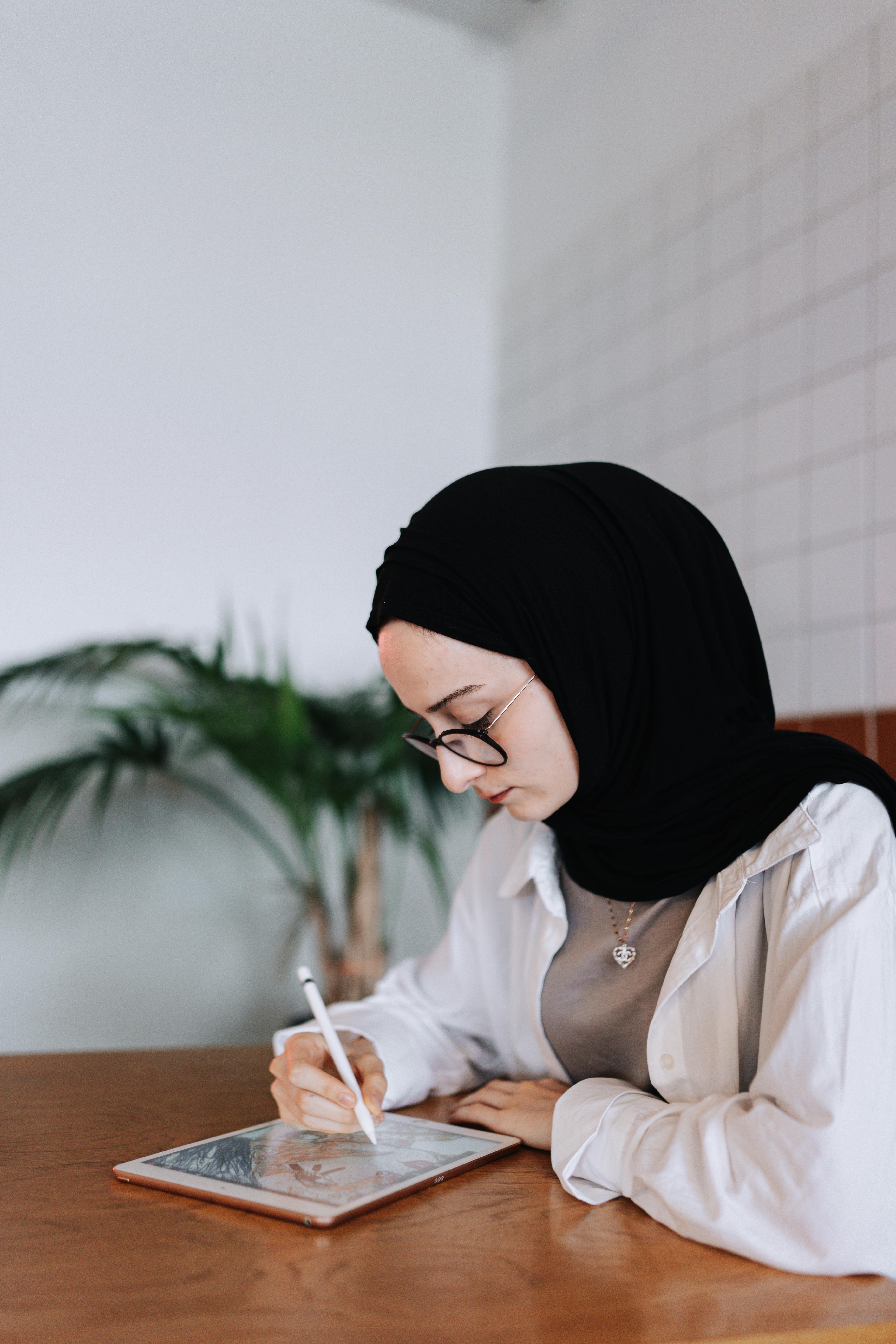 Young woman in hijab drawing on a tablet with a stylus indoors, showcasing creativity and technology.