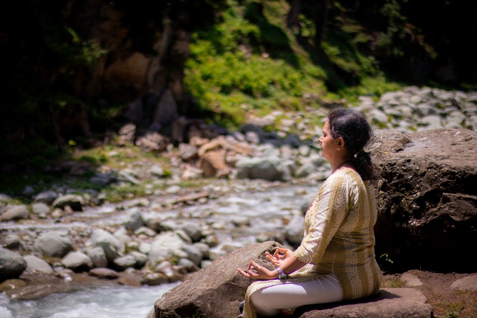 Woman practicing meditation near a tranquil river in a rocky, lush environment. Woman practicing meditation near a tranquil river in a rocky, lush environment.
