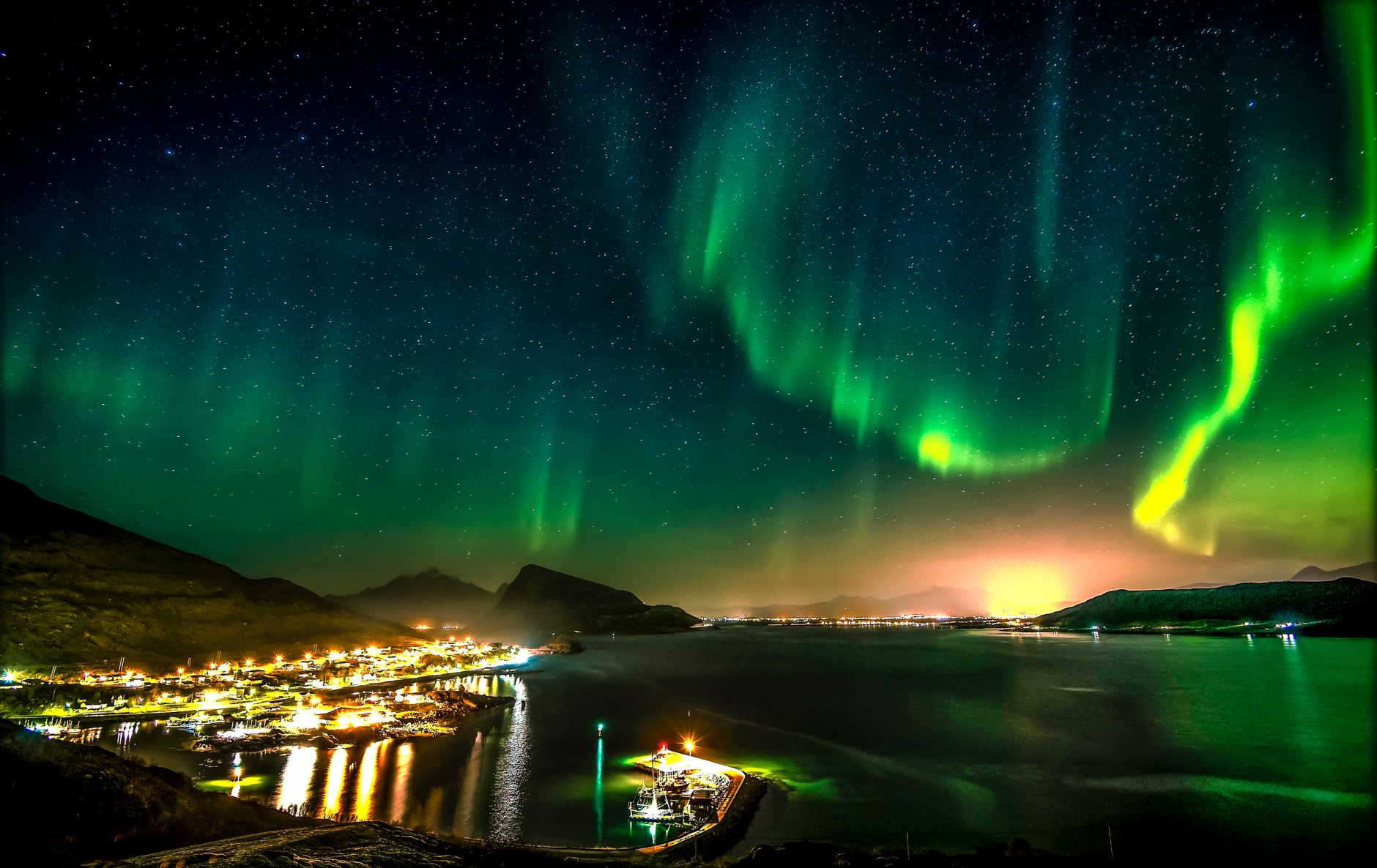 Captivating aurora borealis illuminates Napp, Nordland with vibrant green hues, reflecting over the serene harbor waters at night.