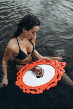 A woman in swimwear holding an ornate mirror while standing in water, reflecting her image.