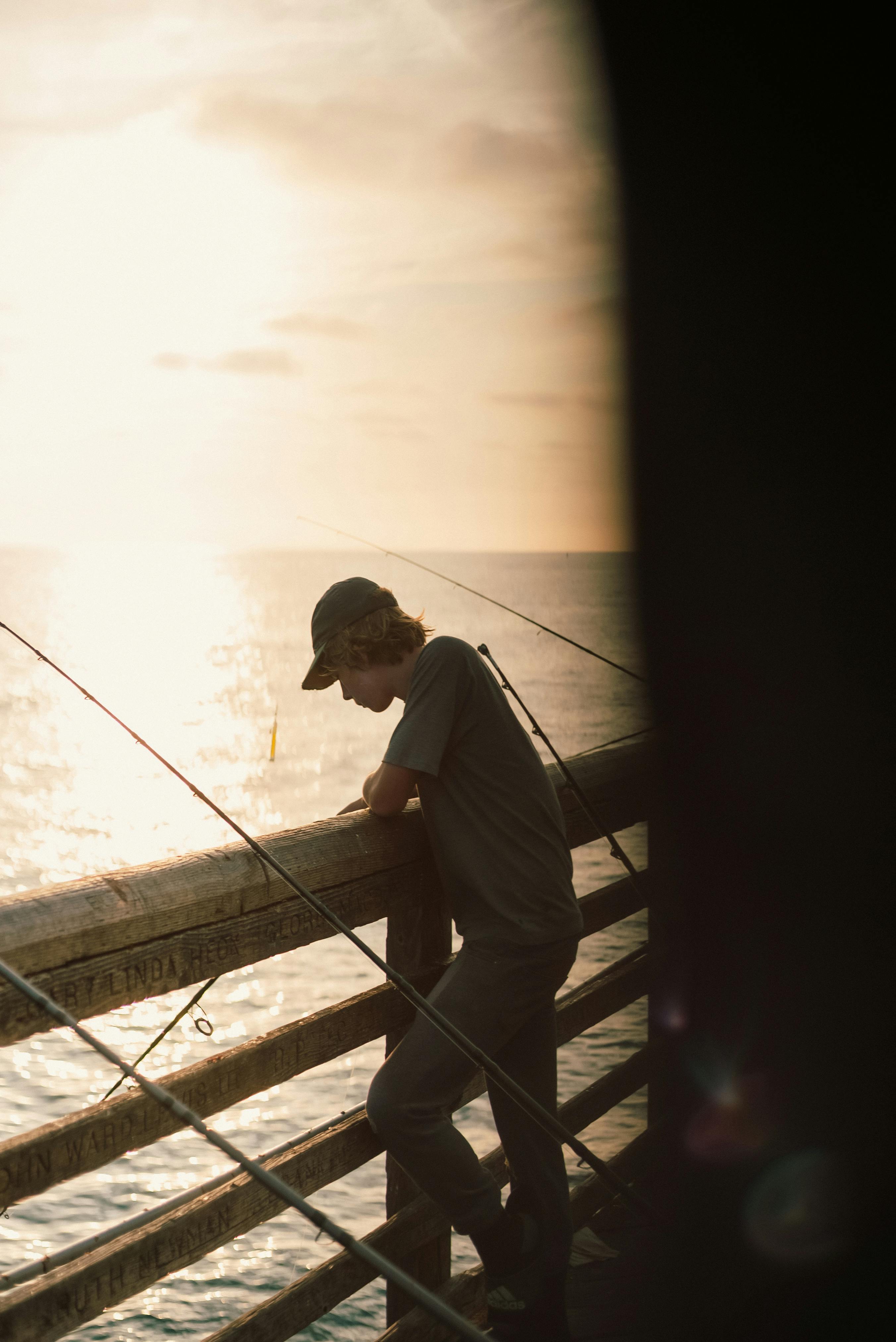 A man leans over a pier railing with fishing rods, silhouetted by the setting sun over the ocean.
