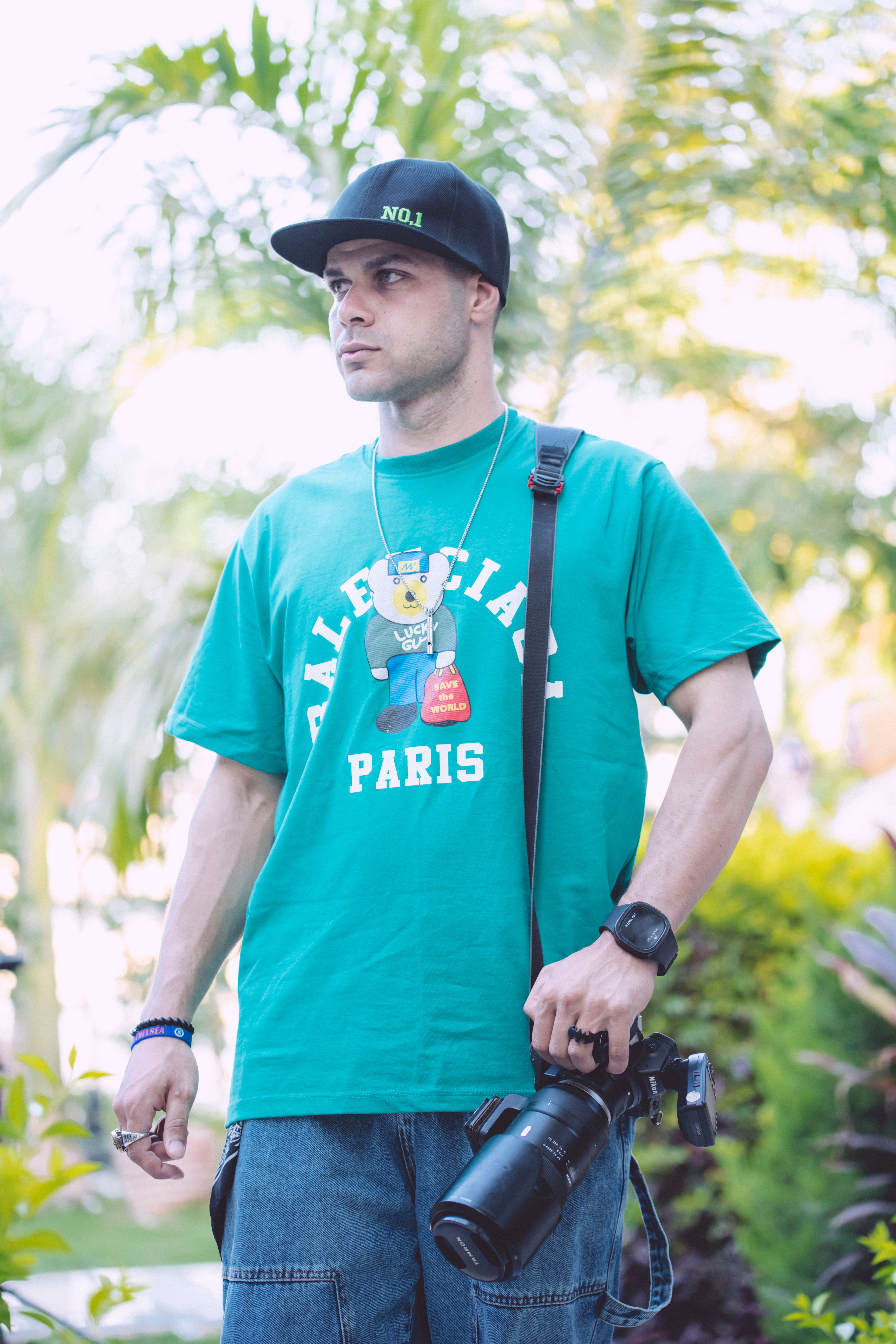 Portrait of a man in a green T-shirt and cap holding a camera outdoors, exuding a casual vibe.