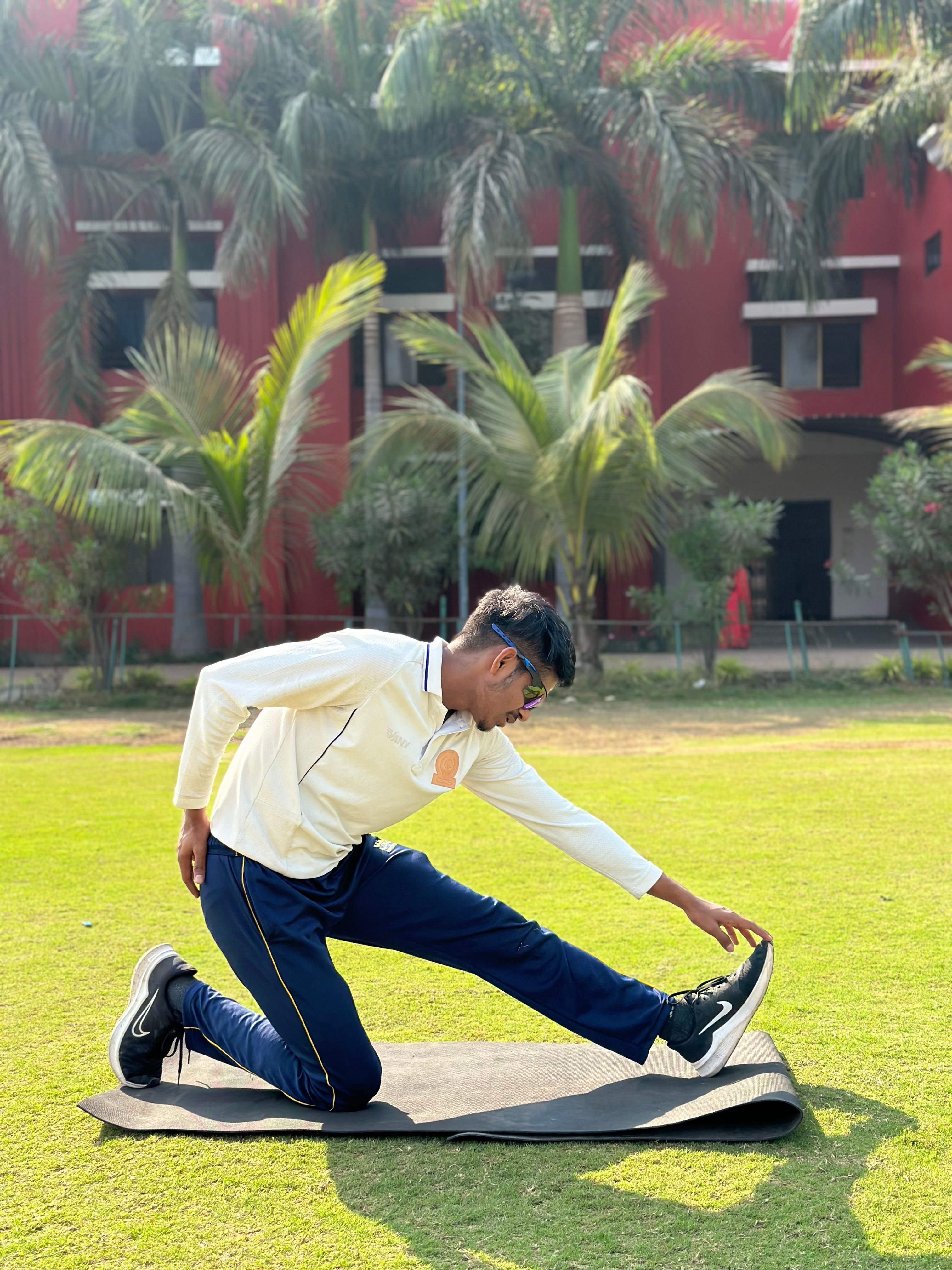 A man stretching in yoga pose on a grass mat outdoors with a building backdrop.