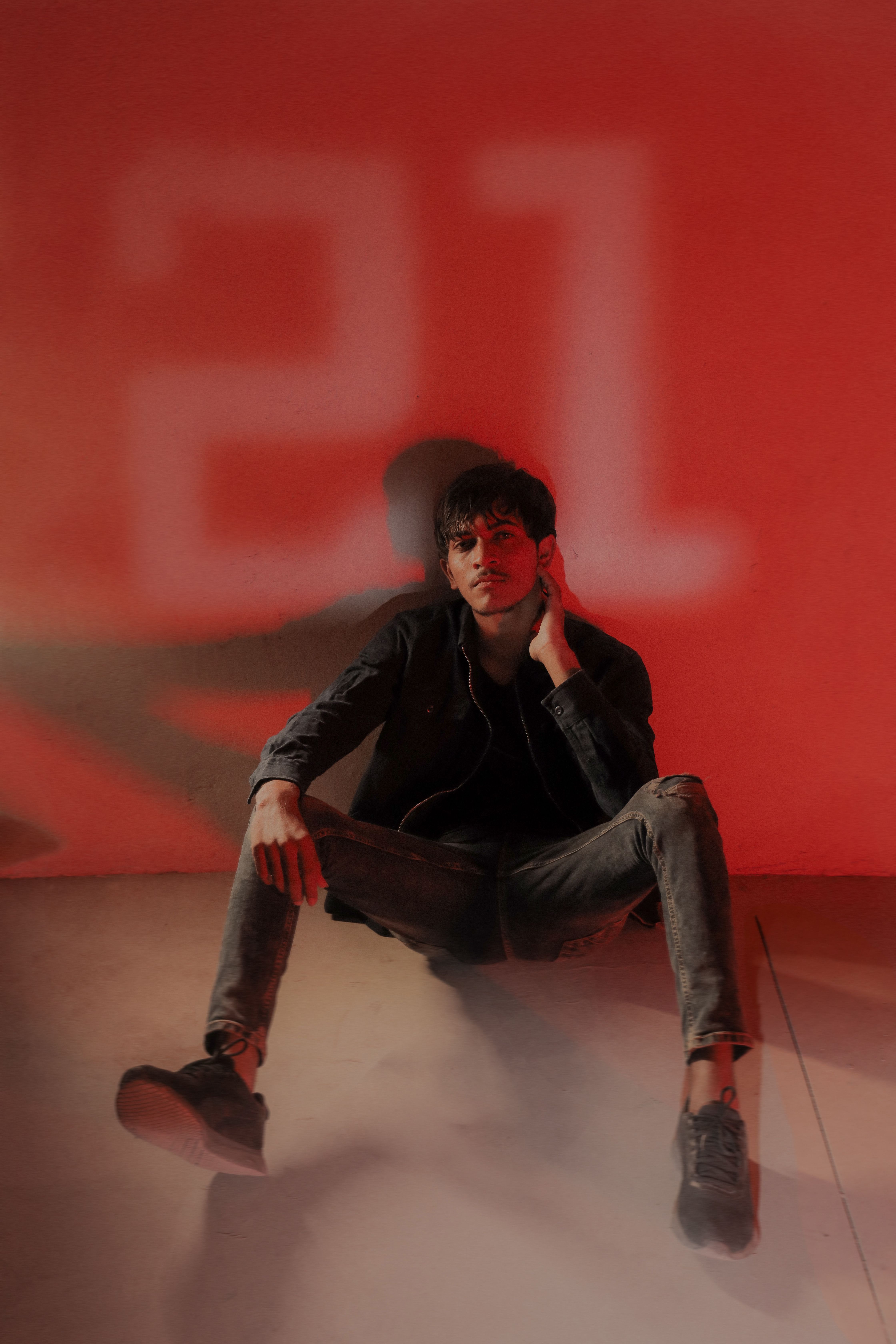 Portrait of a man sitting against a red-lit wall in Morbi, India, depicting casual fashion and moody atmosphere.