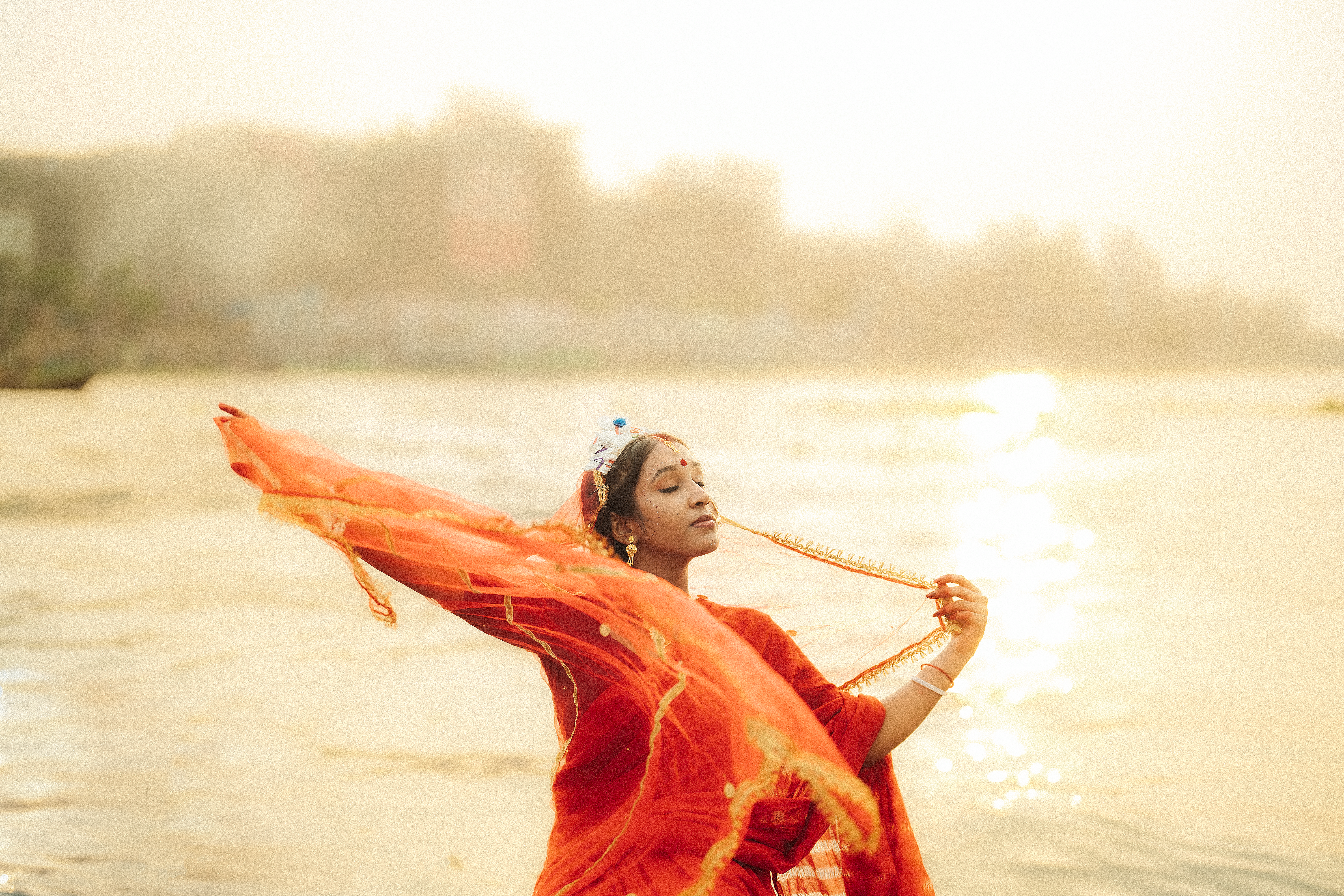 A serene Indian woman gracefully posing in an orange saree by the water during sunset.