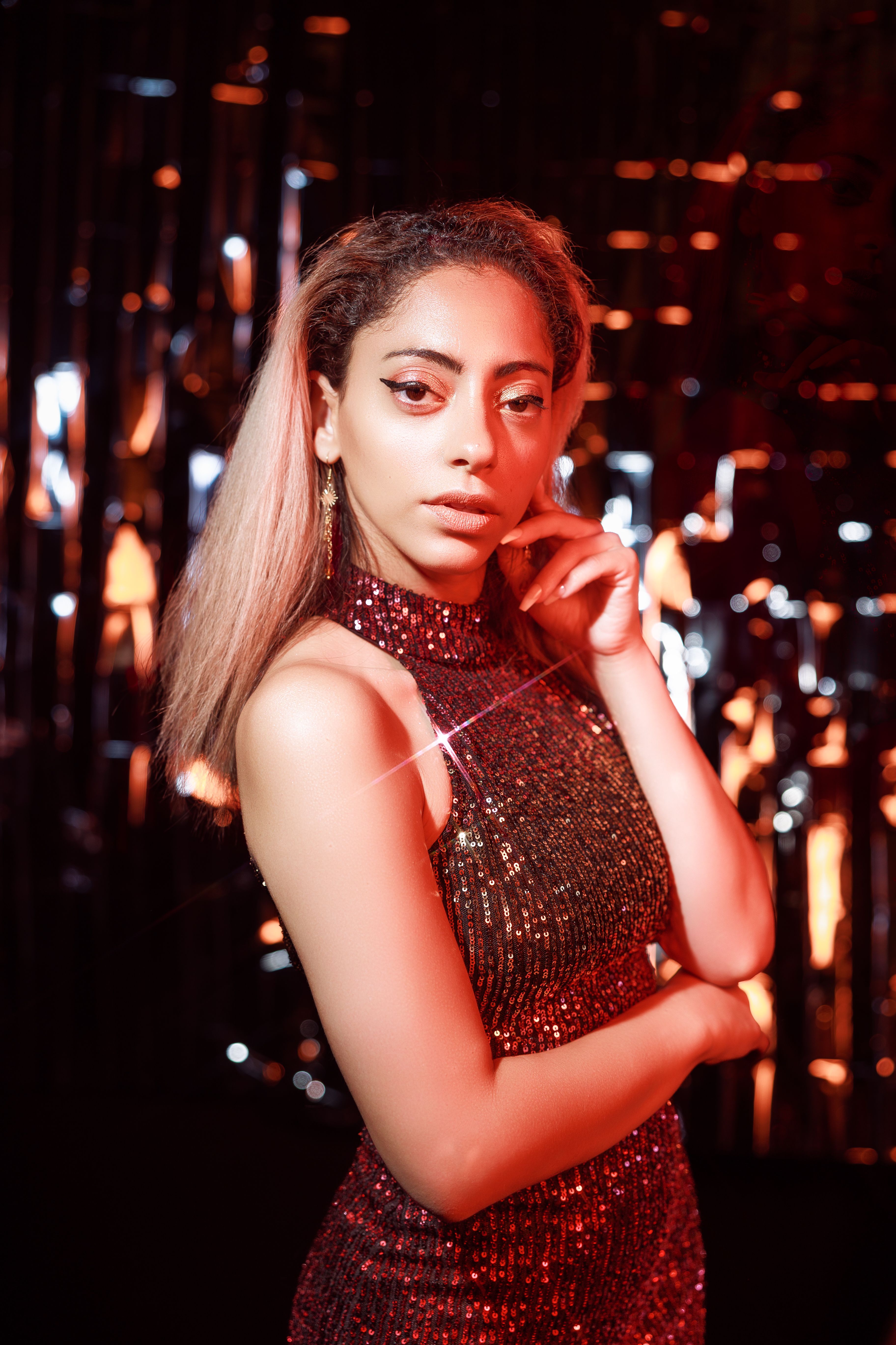 A stylish woman in a glittering dress poses gracefully with dramatic lighting in an indoor setting.