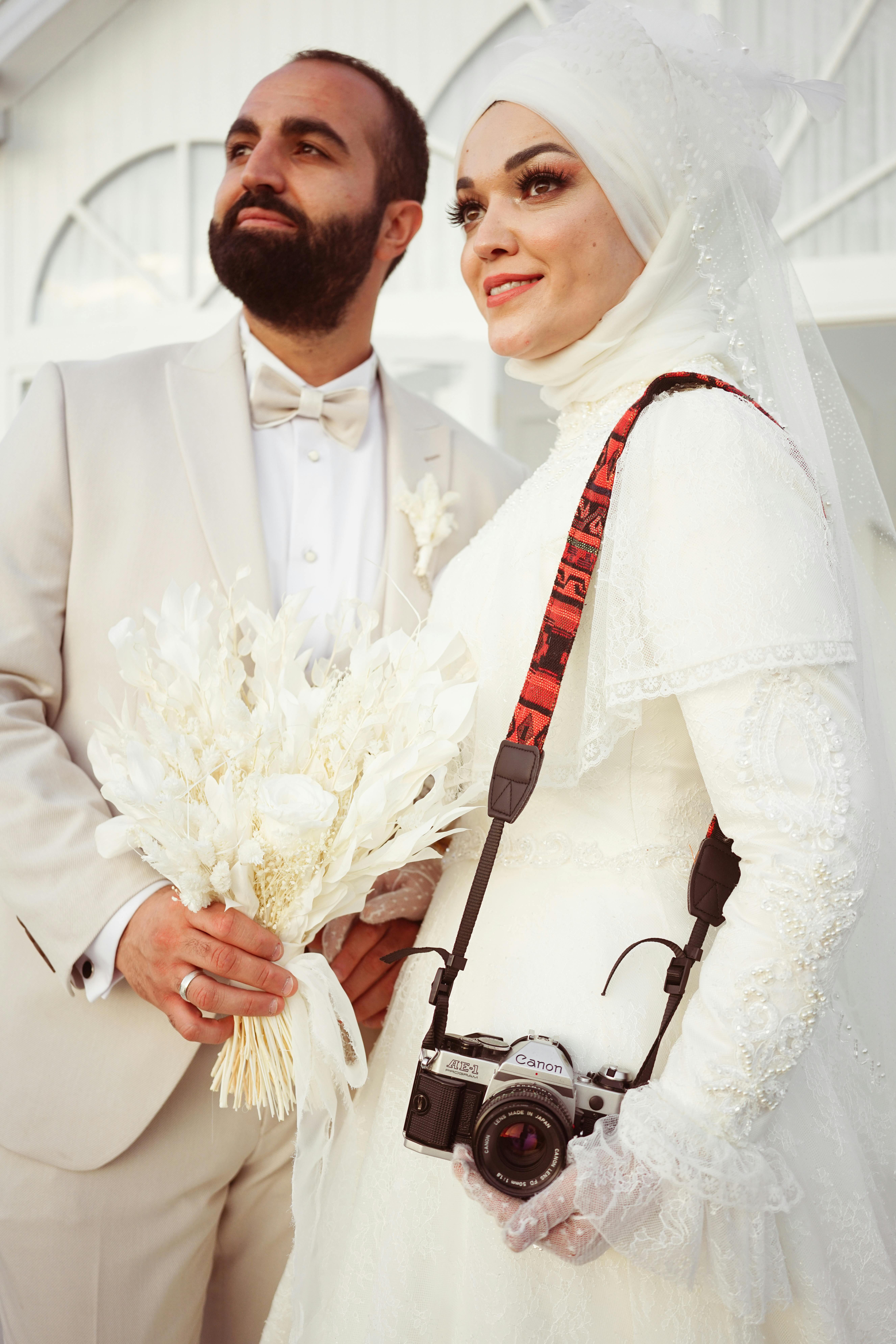 Beautiful wedding portrait of a Muslim couple, bride in hijab holding camera, groom with bouquet.