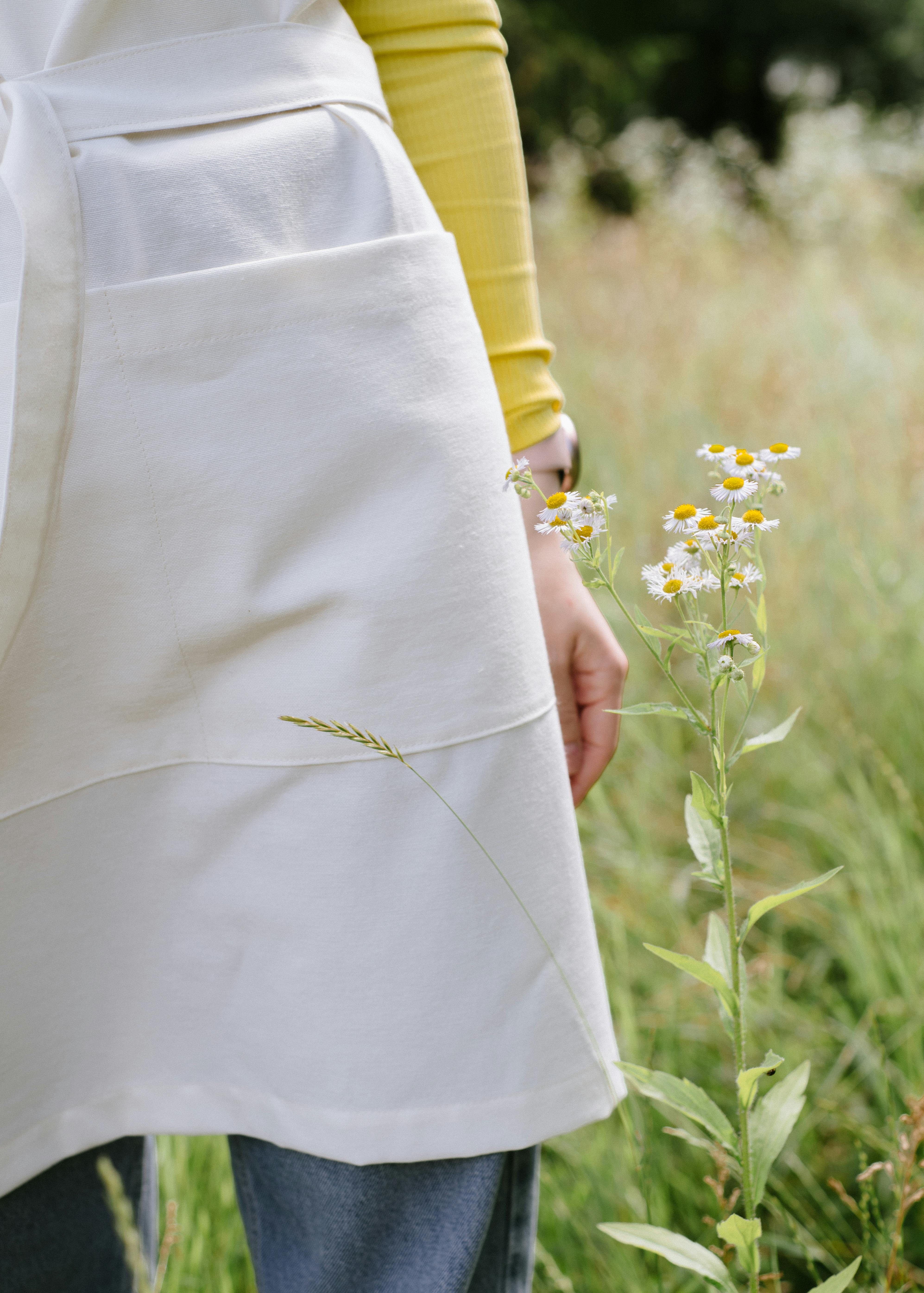 A woman standing in a meadow wearing an apron surrounded by wildflowers on a summer day.