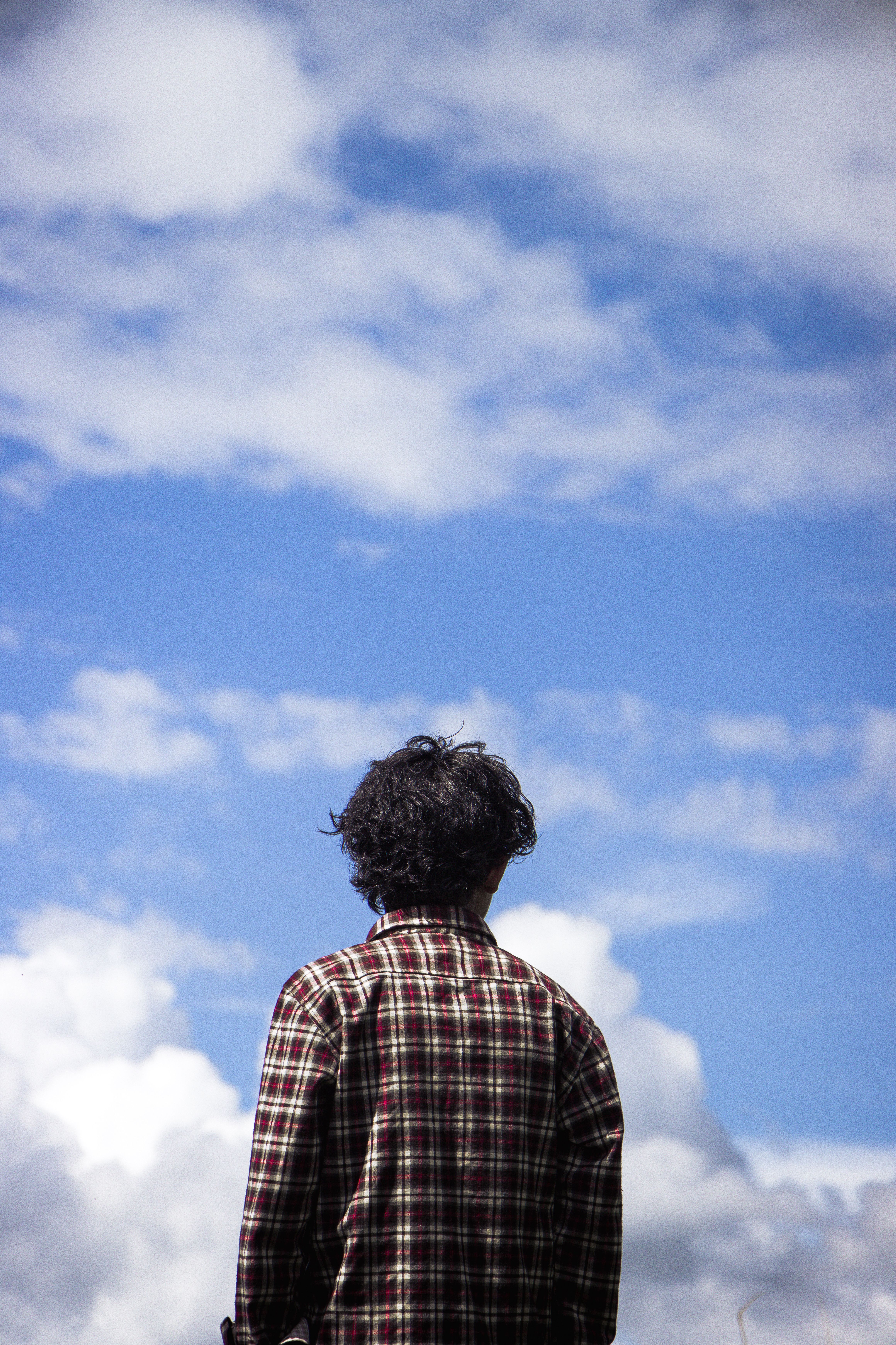 Man with curly hair standing outside, looking at sky with fluffy clouds, creating a thoughtful scene.